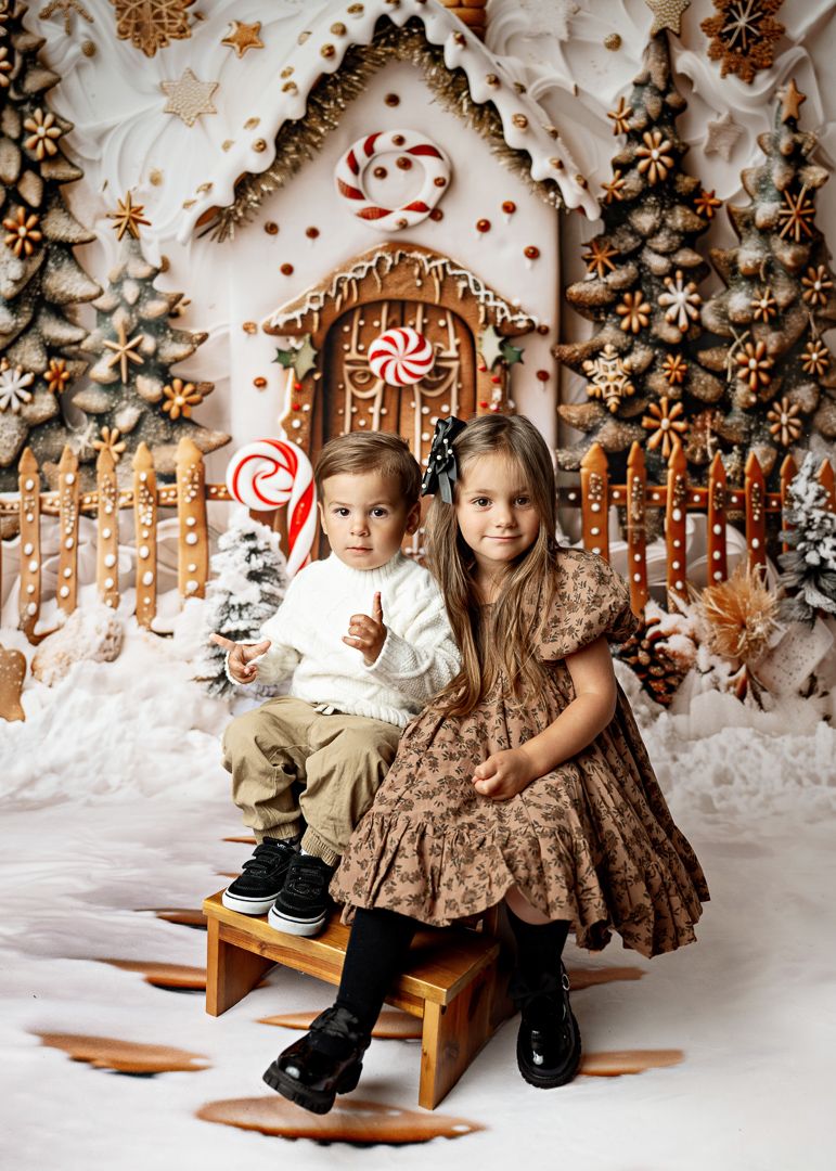 Two children pose in front of a festive gingerbread house backdrop for a Christmas portrait.