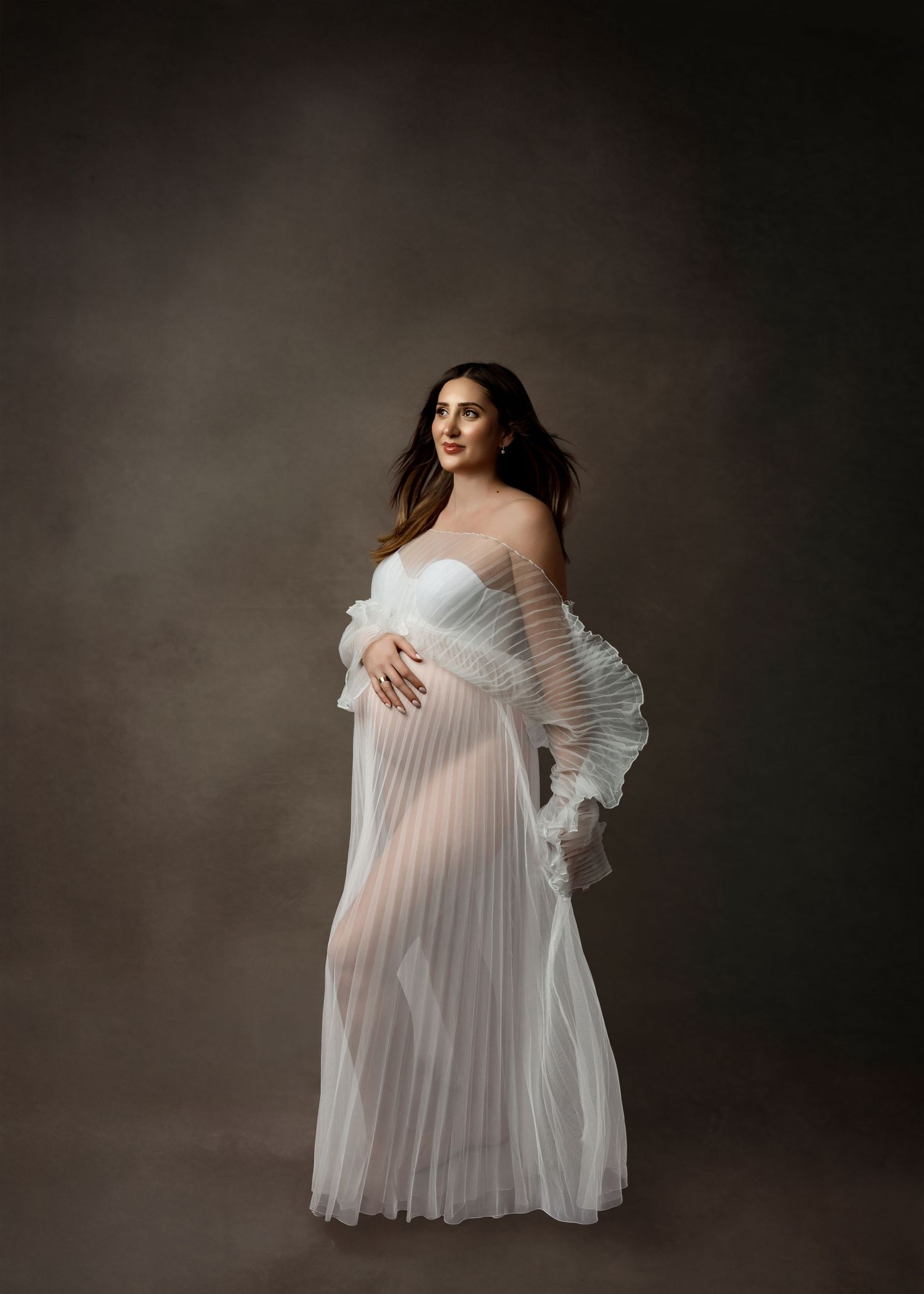 Elegant maternity portrait featuring a flowing white pleated gown with off-shoulder ruffled sleeves against a dark backdrop.