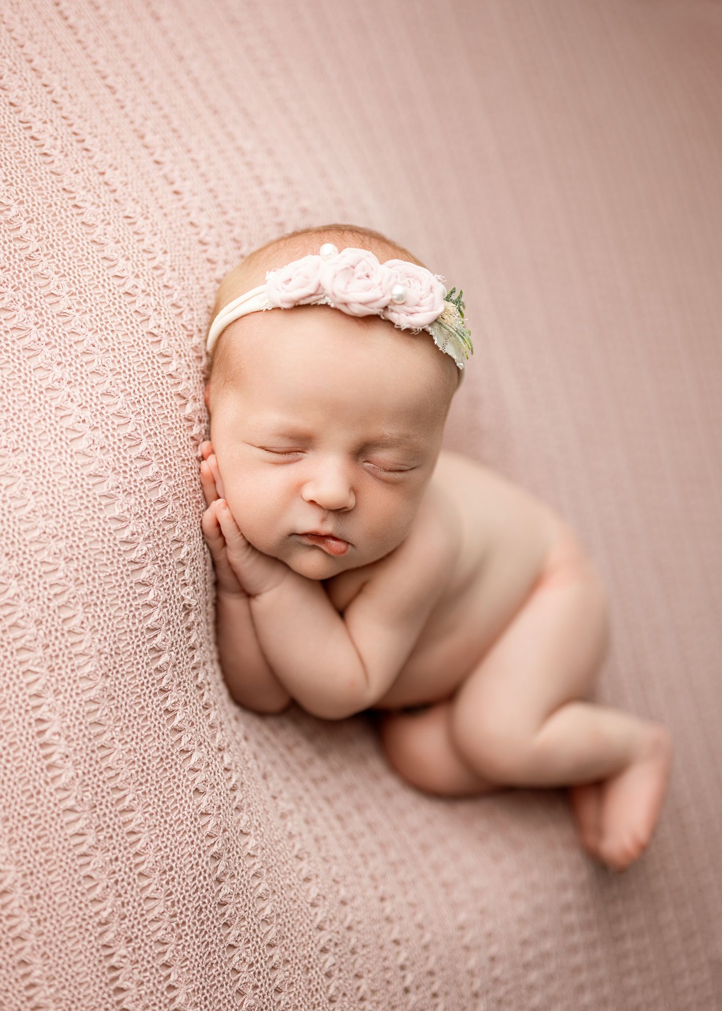 Sleeping newborn baby curled up on soft pink knit blanket wearing delicate floral headband in peaceful studio portrait.