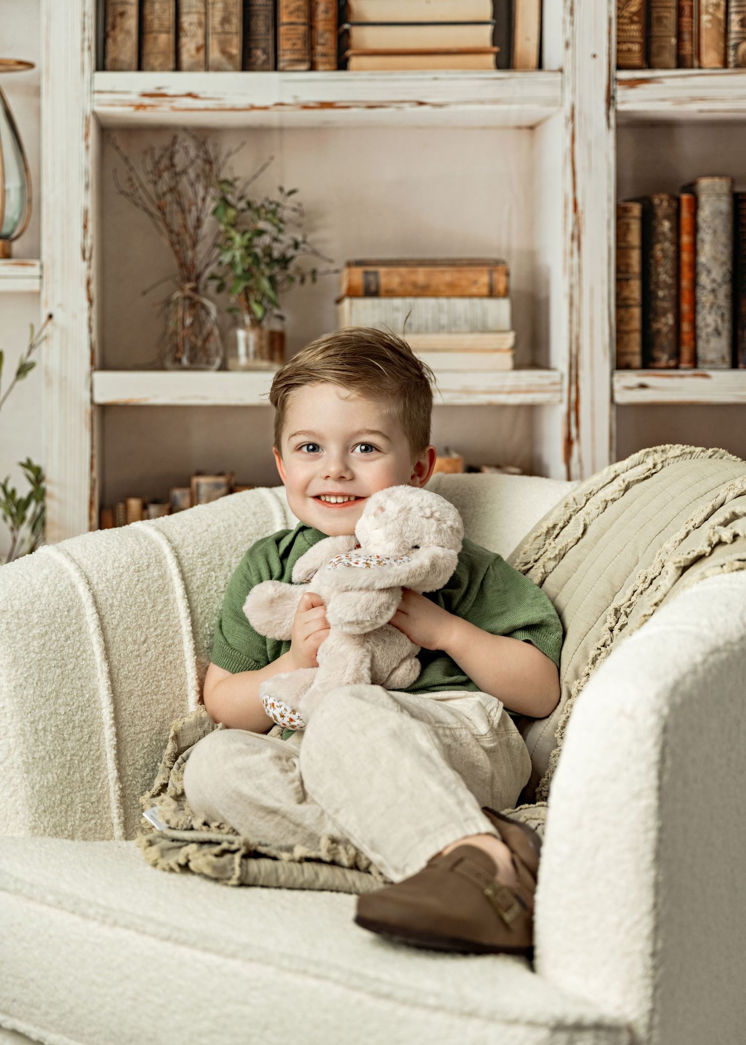 A cozy reading nook features a white armchair, vintage bookshelves, and a soft plush toy against a warm neutral backdrop.