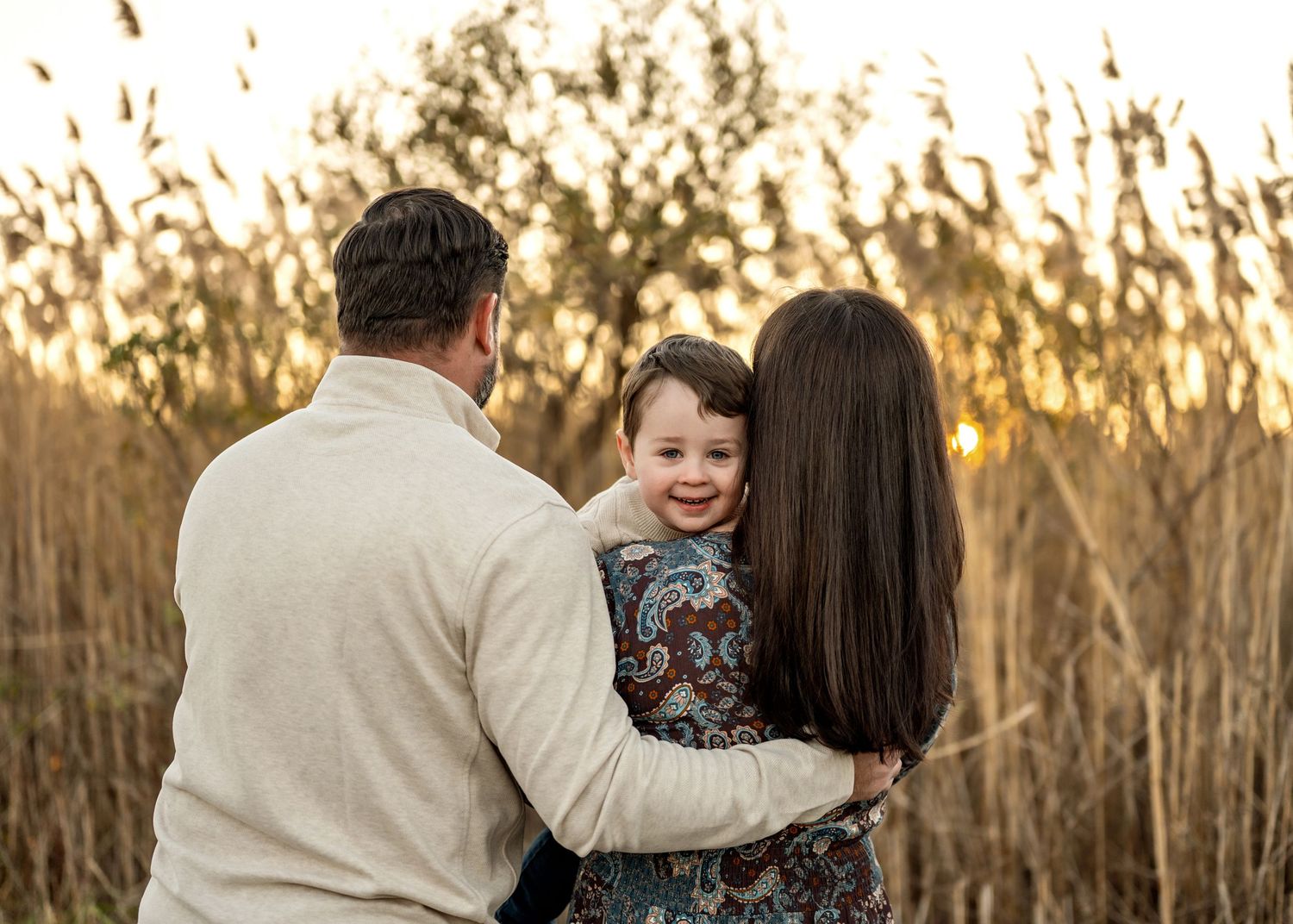 A family embraces outdoors during golden hour with tall trees silhouetted in the background.