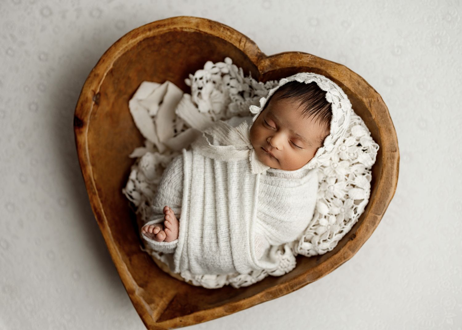 Sleeping newborn swaddled in white fabric rests peacefully in wooden heart-shaped bowl with lace backdrop.