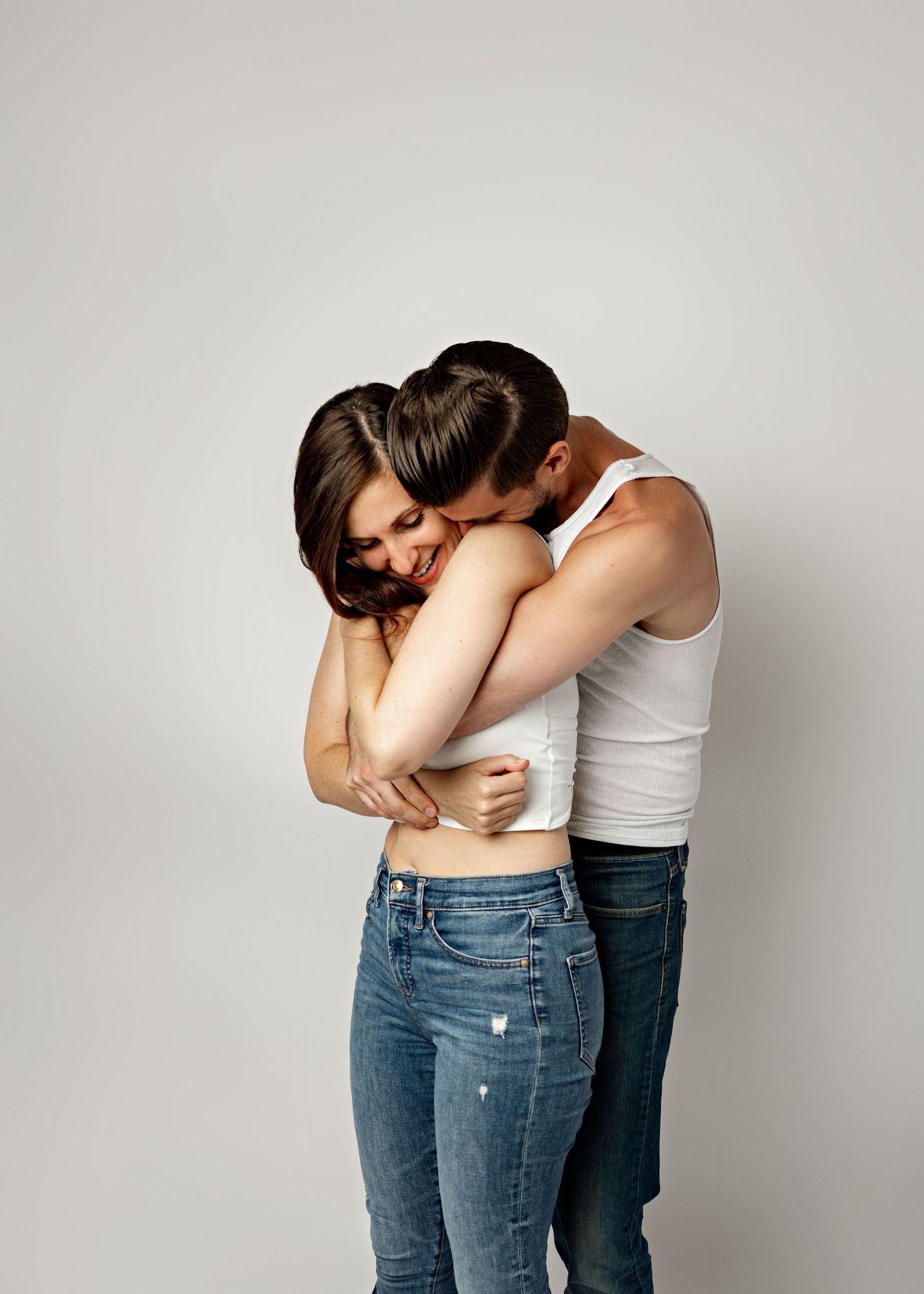 Intimate couple wearing jeans and white tops embrace tenderly against a light gray background.