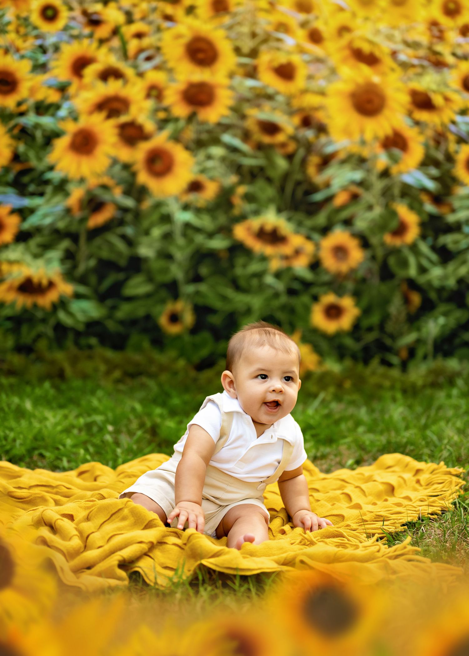A joyful baby sits on a yellow blanket surrounded by a vibrant field of blooming sunflowers.