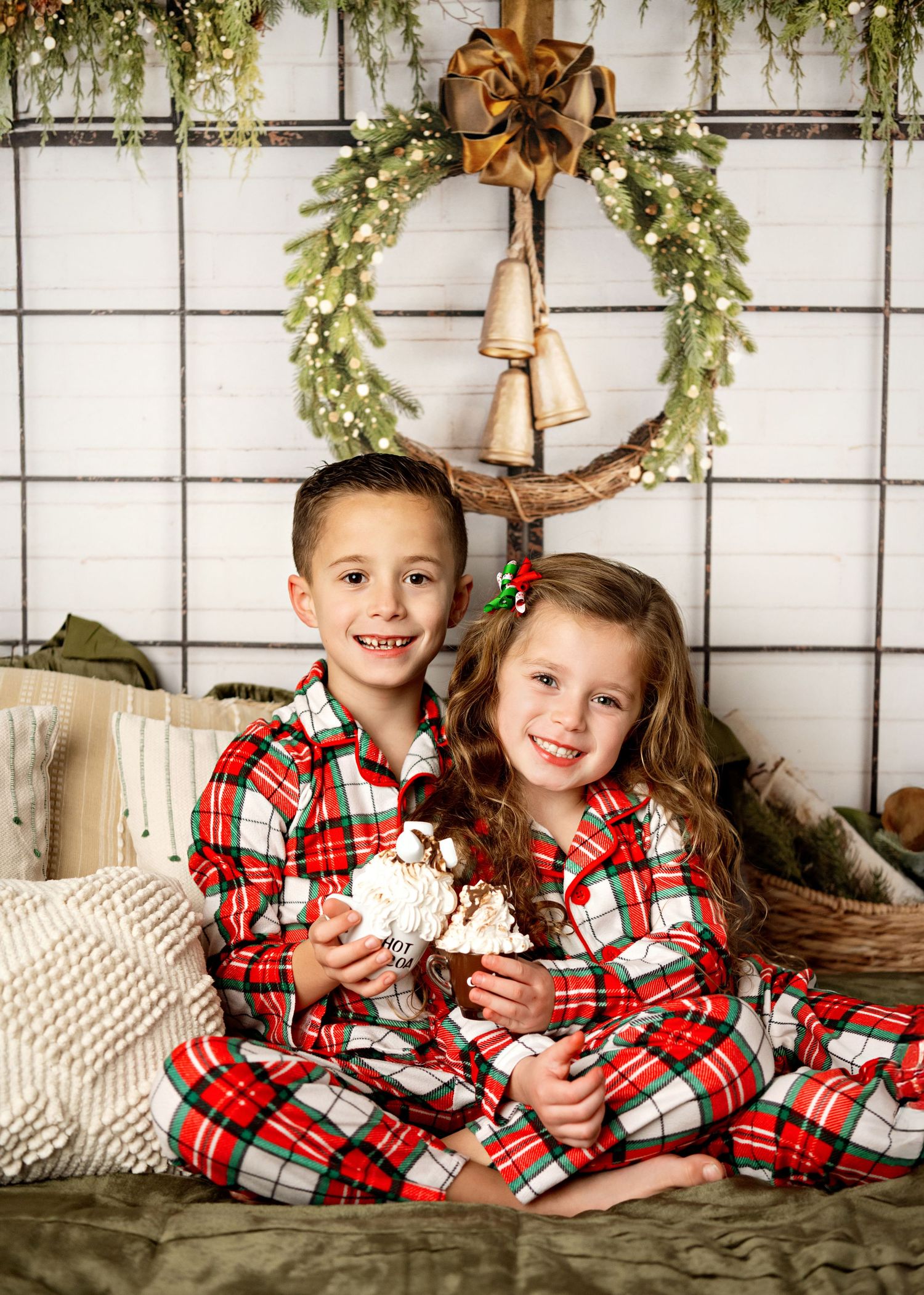 Two children in matching red plaid Christmas pajamas sit together on a couch beneath a festive holiday wreath.