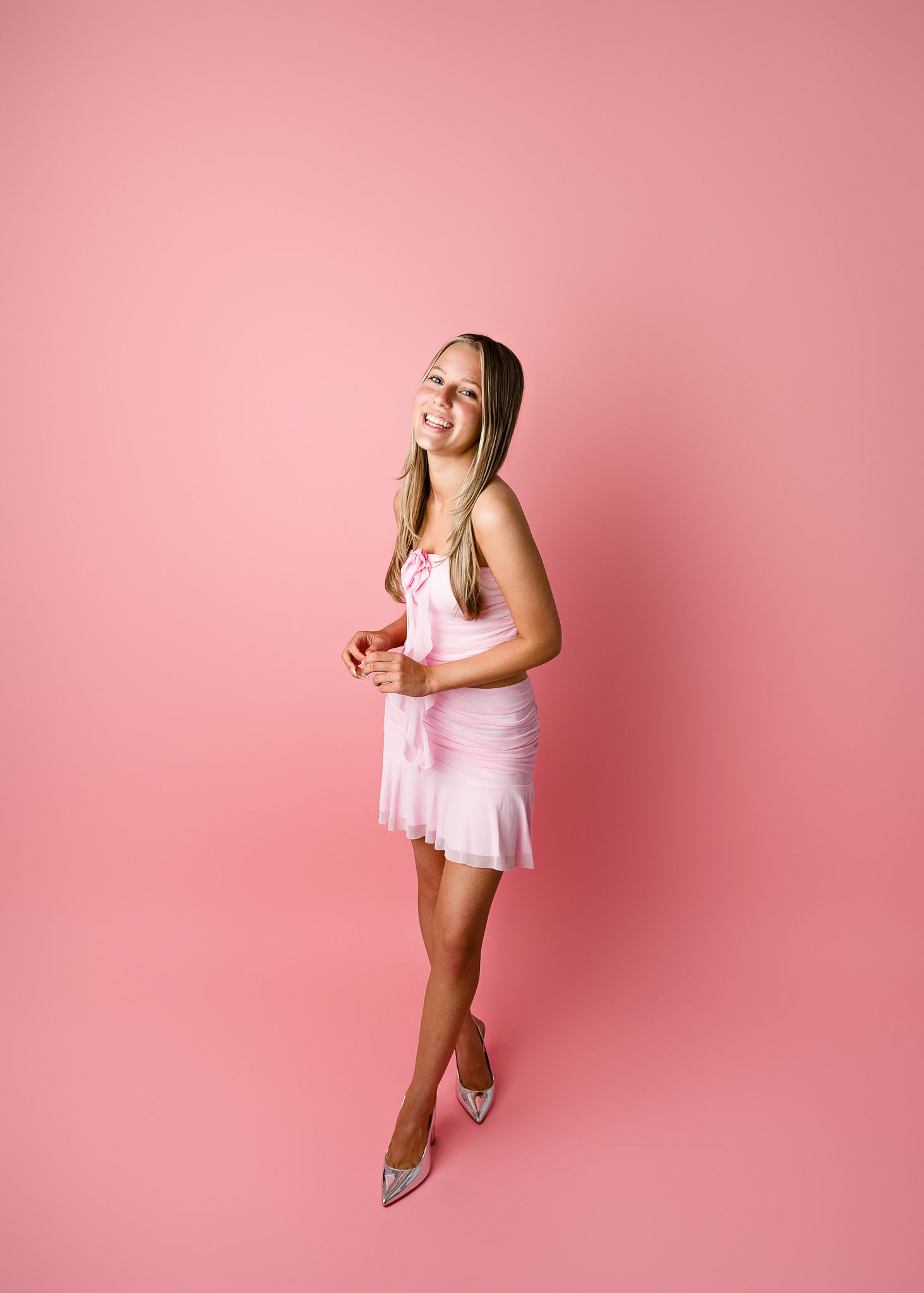 Elegant model in pale pink mini dress and heels stands smiling against rosy pink background.