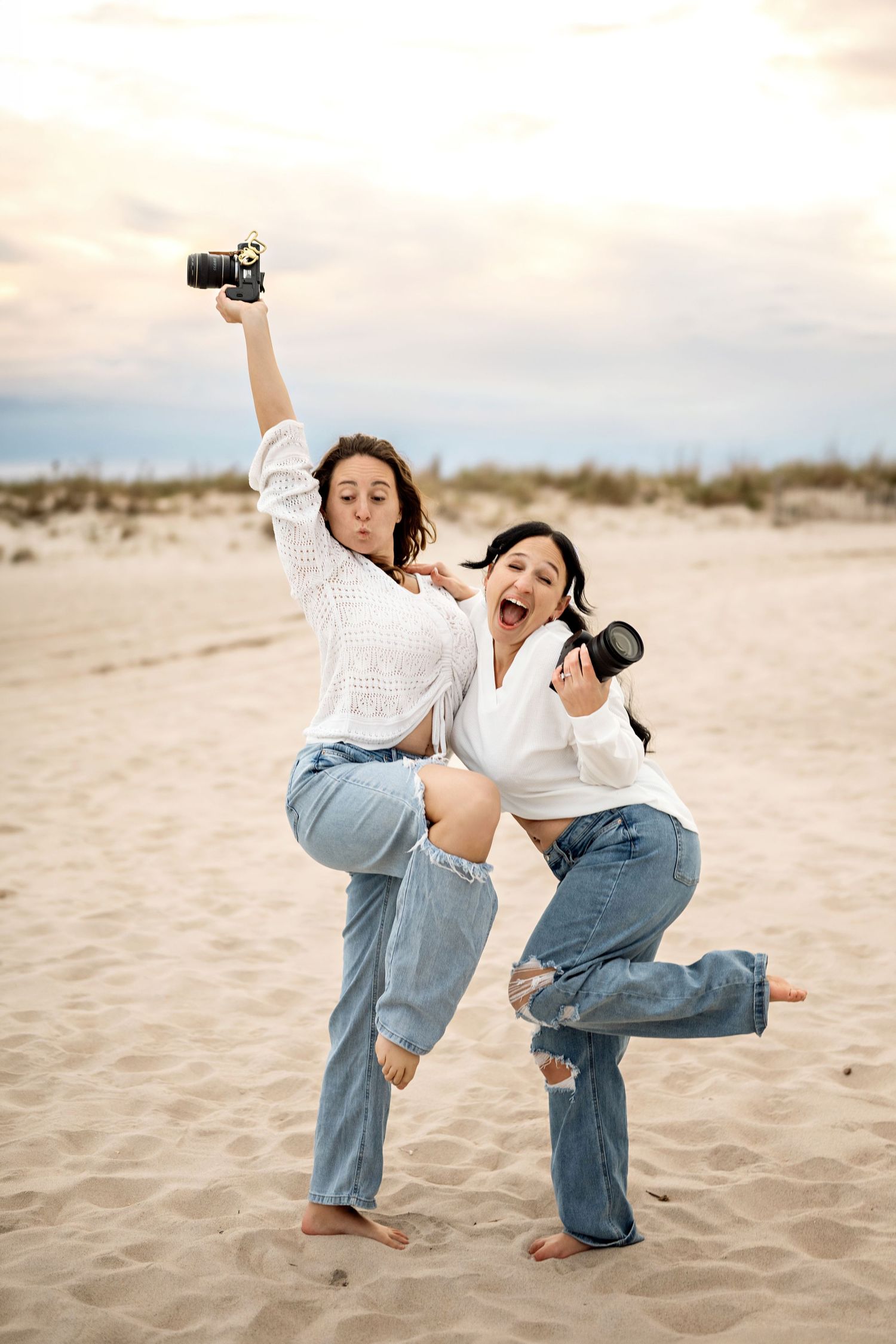 Two women in white shirts and jeans pose playfully on a sandy beach, each holding a camera.