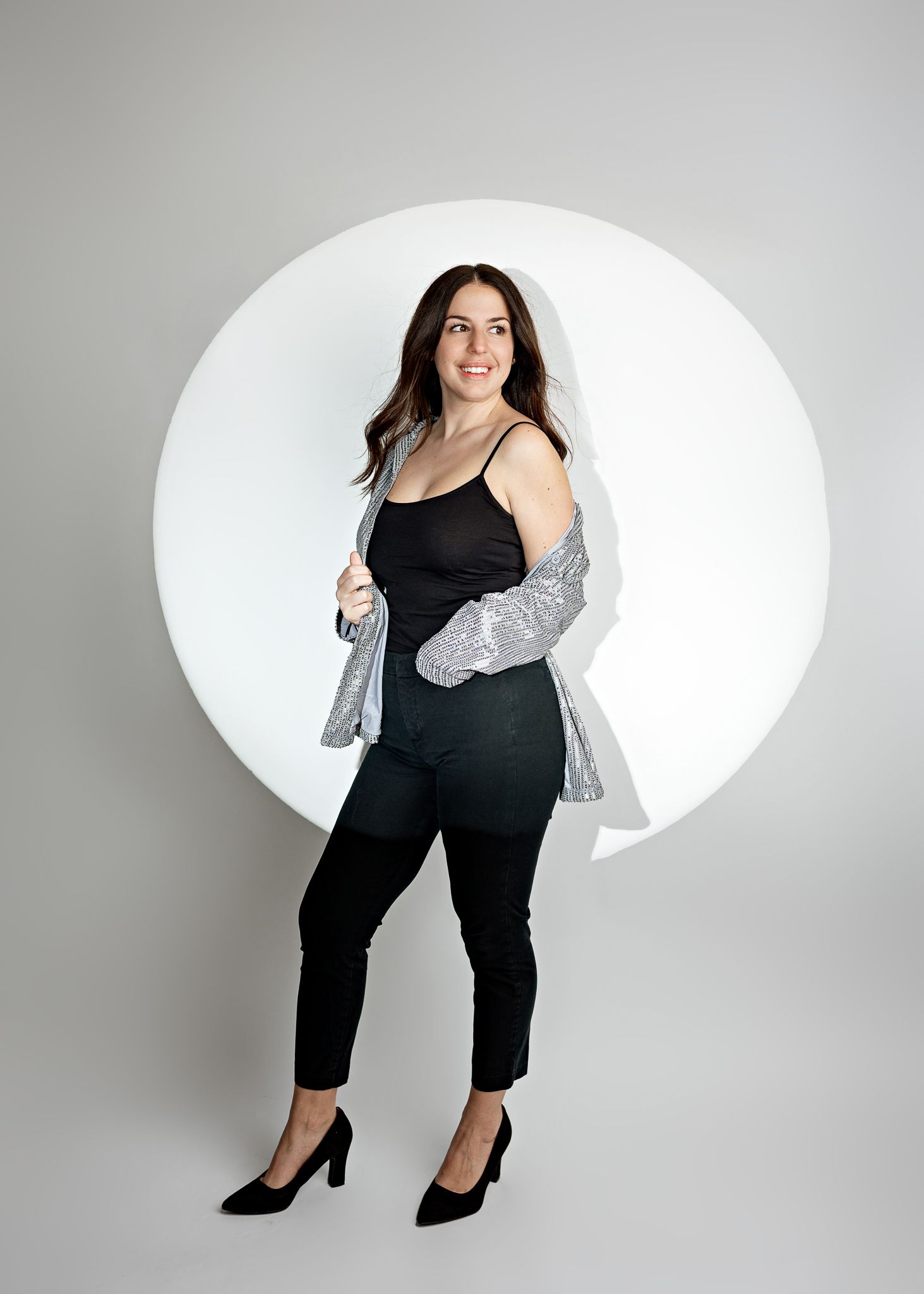 Woman in black outfit poses on gray background with white circular backdrop wearing stilettos and silver cardigan.