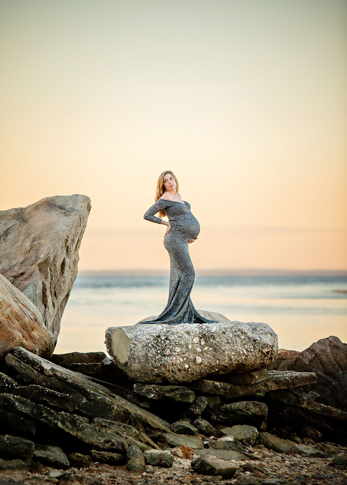 An expectant mother in a flowing gown poses gracefully on coastal rocks during golden hour sunset.
