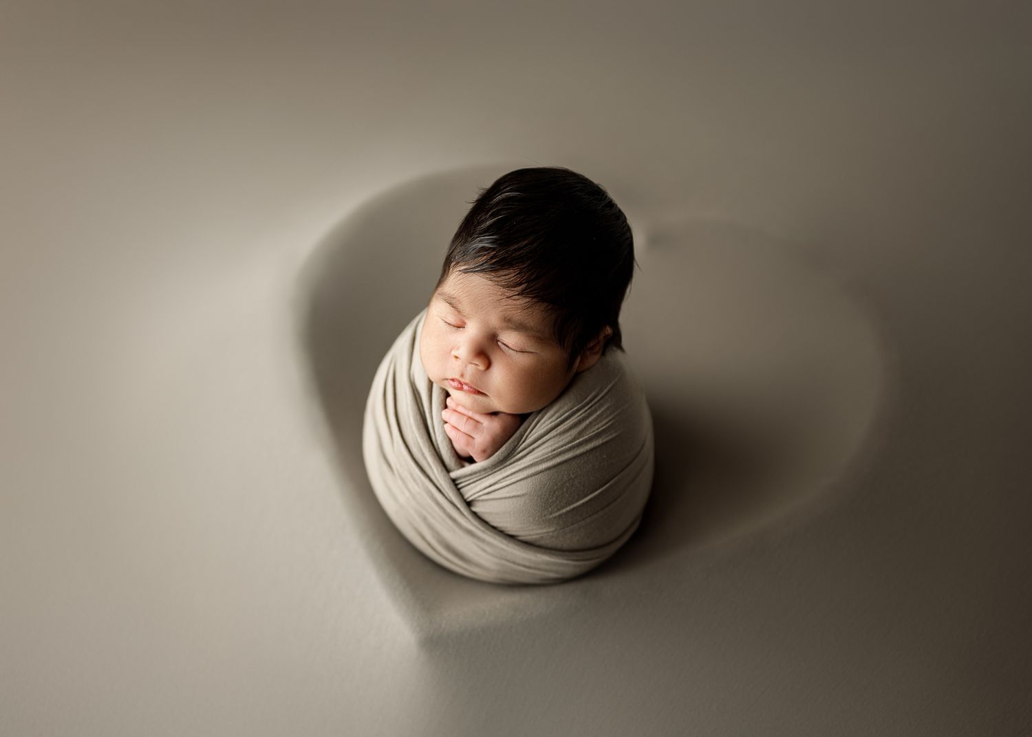 Newborn baby peacefully sleeping wrapped in cream fabric against neutral backdrop with soft shadows.