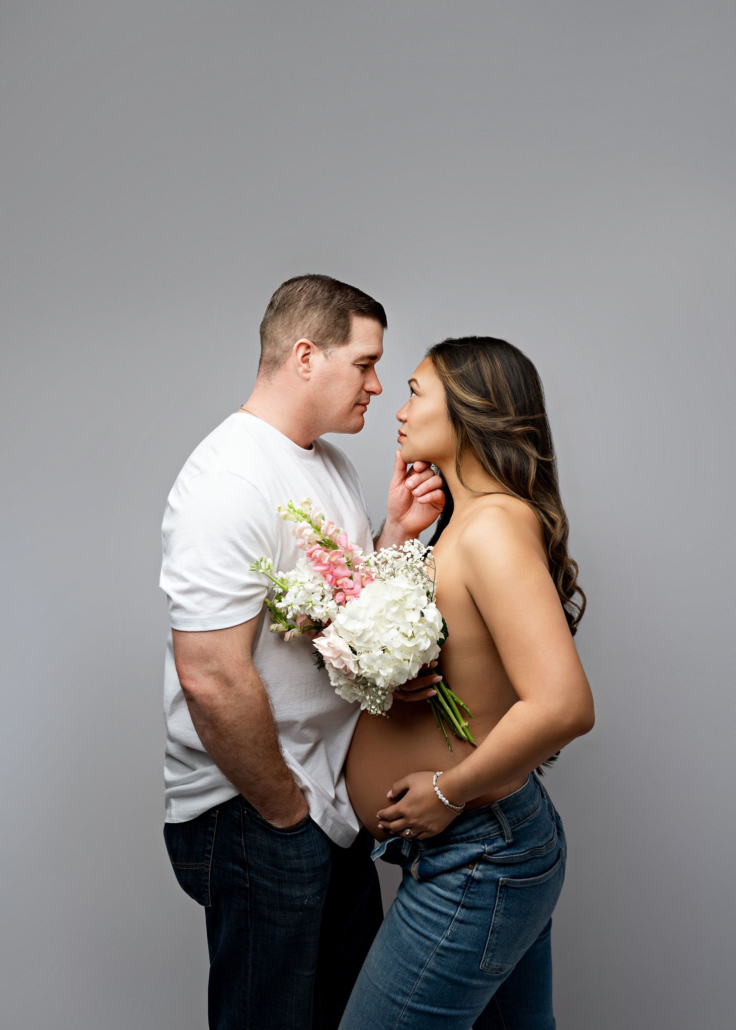 A couple in casual attire share an intimate moment while holding a bouquet of white and pink flowers against a gray backdrop.