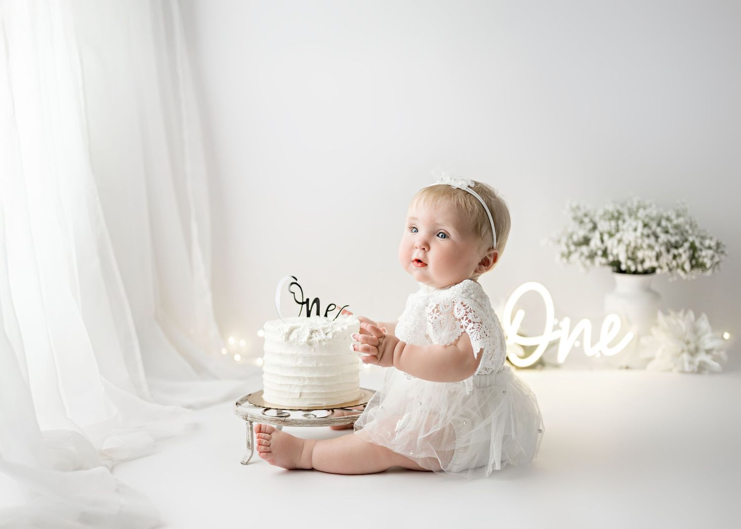 Baby in white dress sits by elegant white cake with neon 'One' sign and flowers in bright ethereal setting.