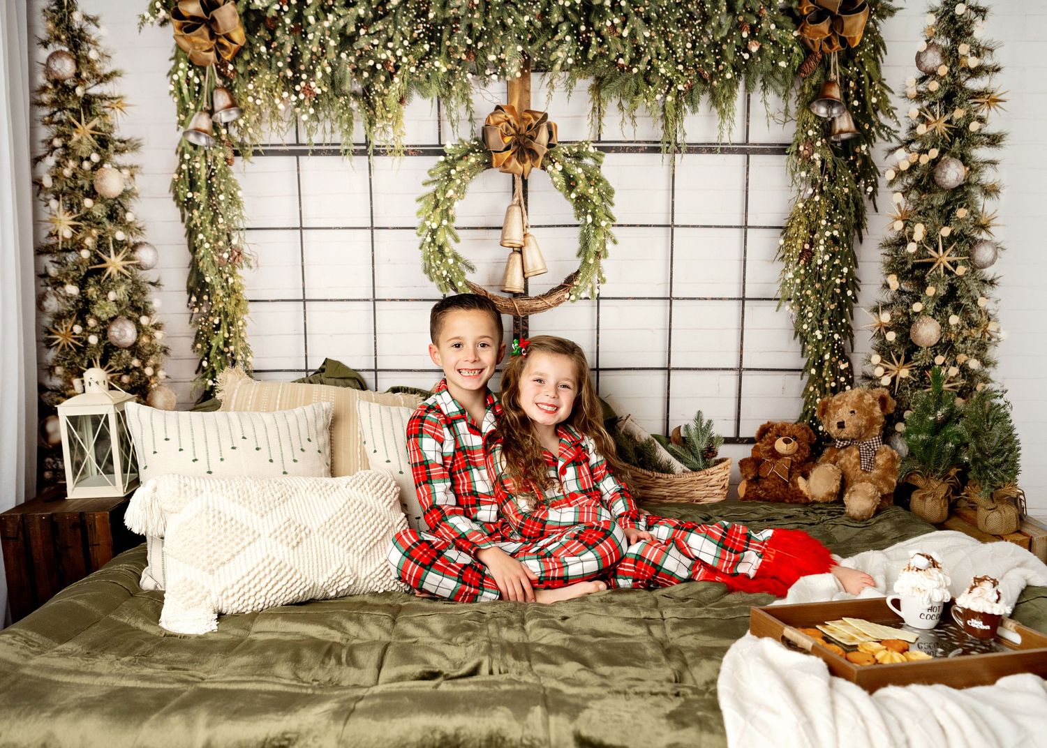 Two people in matching plaid pajamas sit on a bed decorated for Christmas with greenery and lights in a cozy holiday setting.