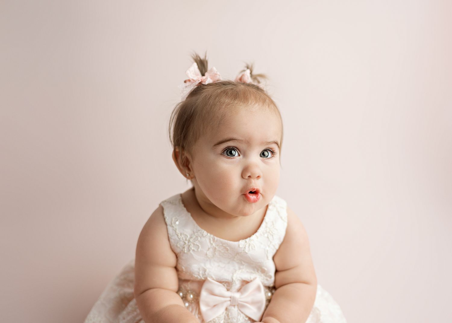 A small child in a white lace dress with a bow sits against a pink backdrop.