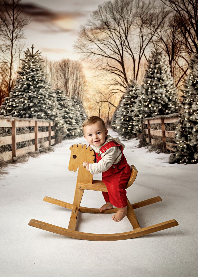 Smiling baby in red overalls on a wooden rocking horse in a snowy Christmas tree farm setting.
