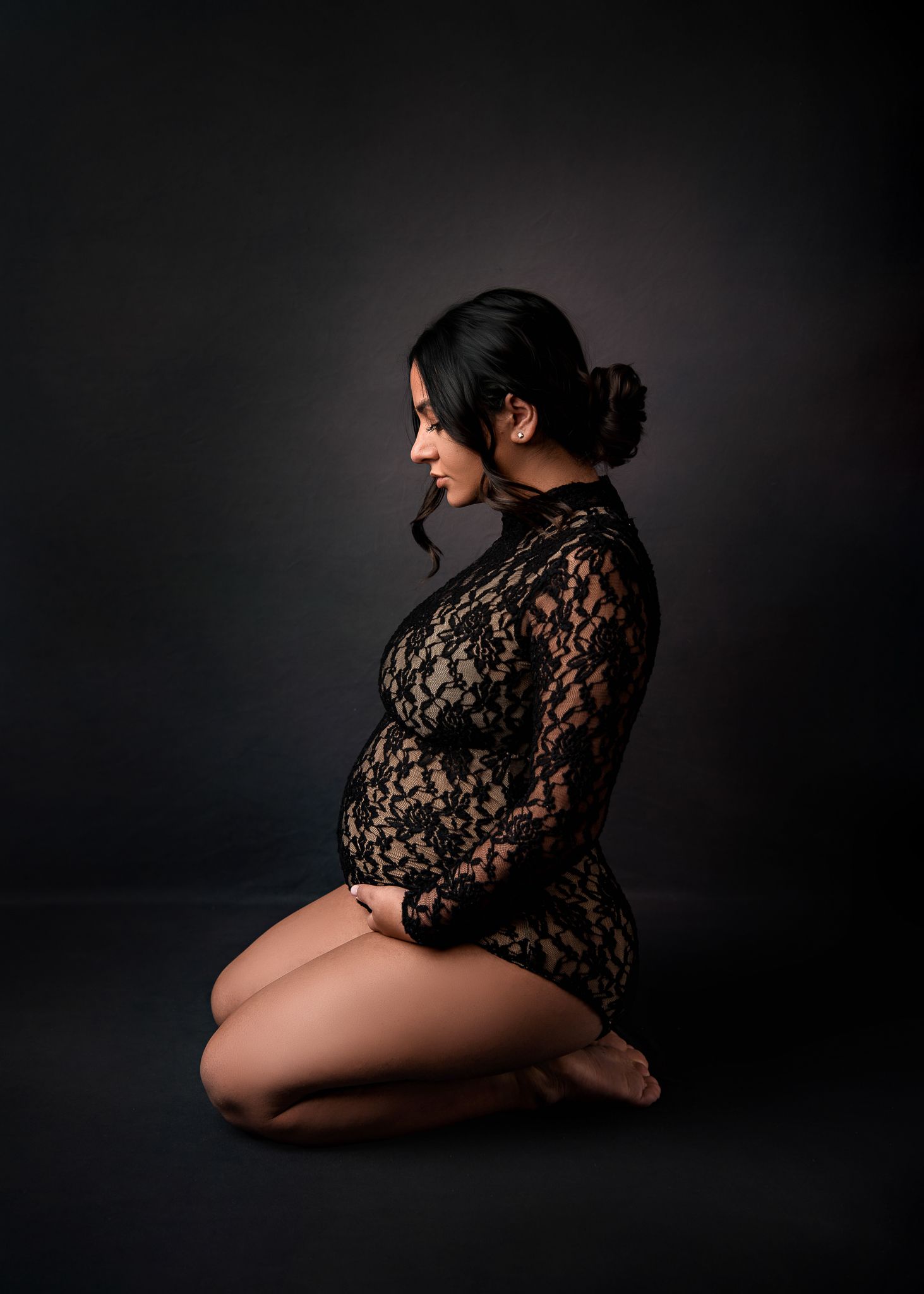 Artistic maternity photo in black lace against dark background with dramatic side lighting creating moody atmosphere.