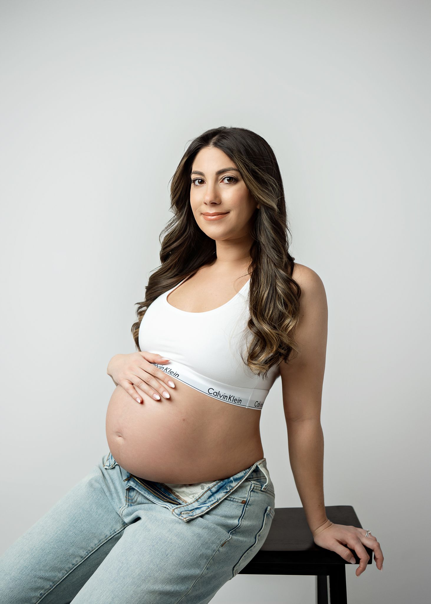 A pregnant woman in a white Calvin Klein sports bra and jeans poses against a light gray background.