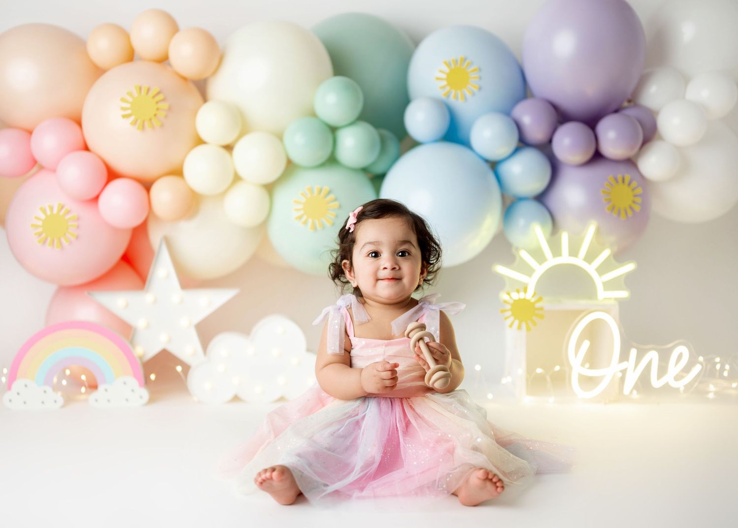 Pastel balloon garland decorates first birthday photo setup with neon lights against white backdrop.