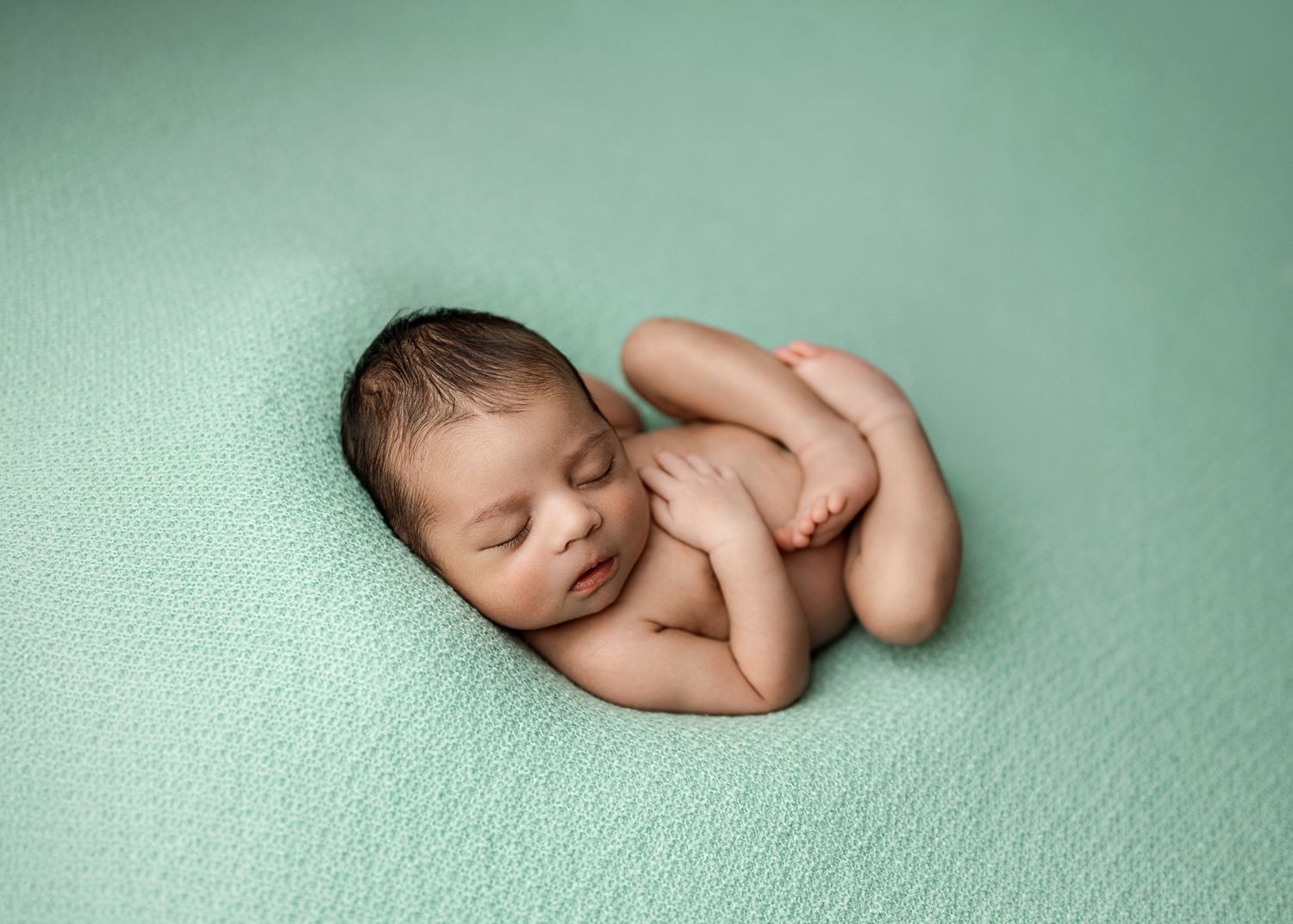 Peaceful sleeping newborn baby curled up on soft mint green blanket in studio lighting.