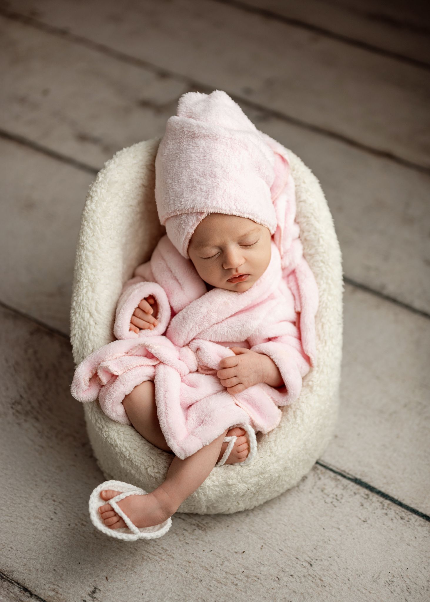 A sleeping newborn in pink swaddle blanket rests peacefully in a white cushioned nest on wooden floor.