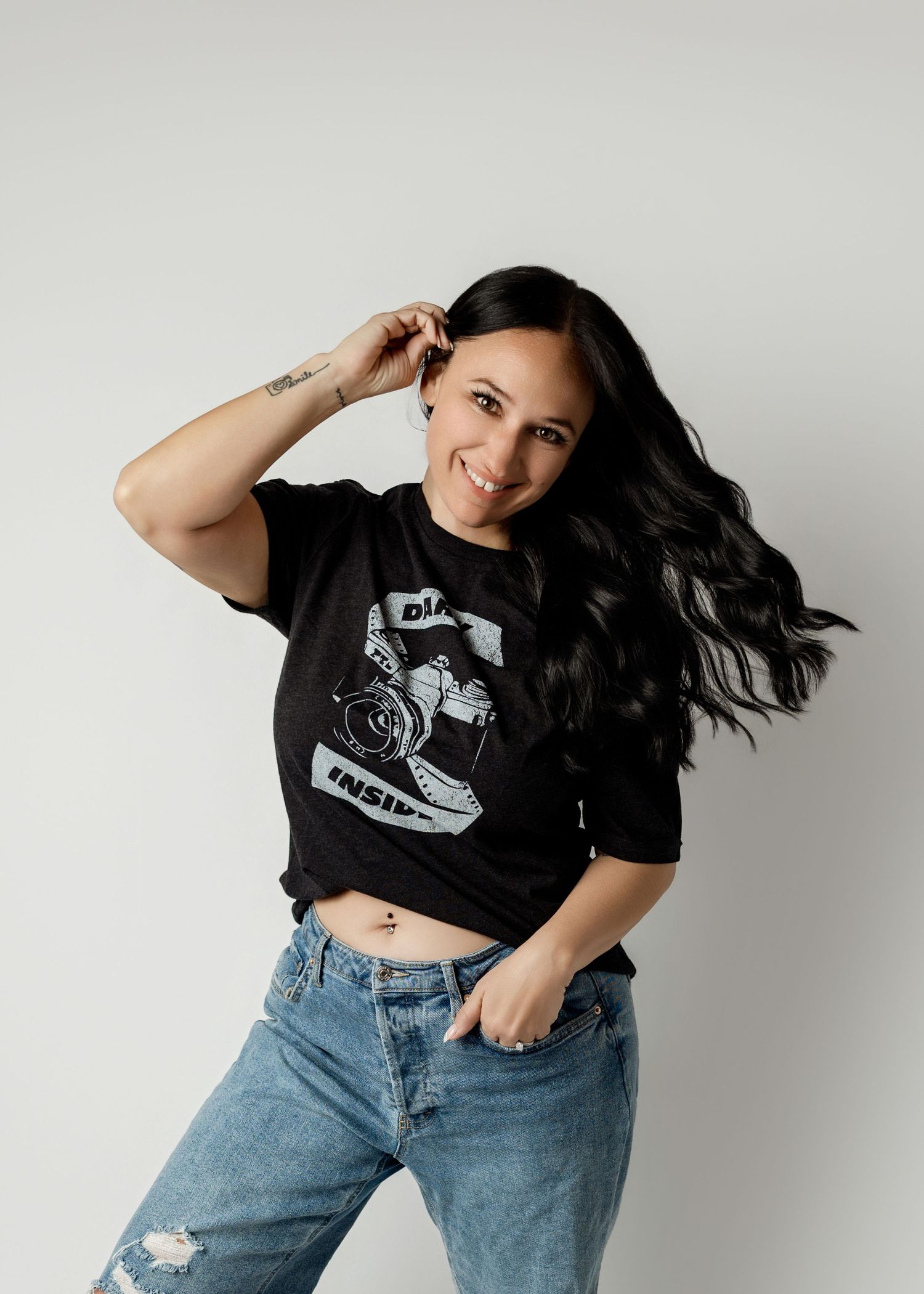 Smiling woman with long dark hair wearing a black graphic tee and ripped jeans, posing confidently against a white background.