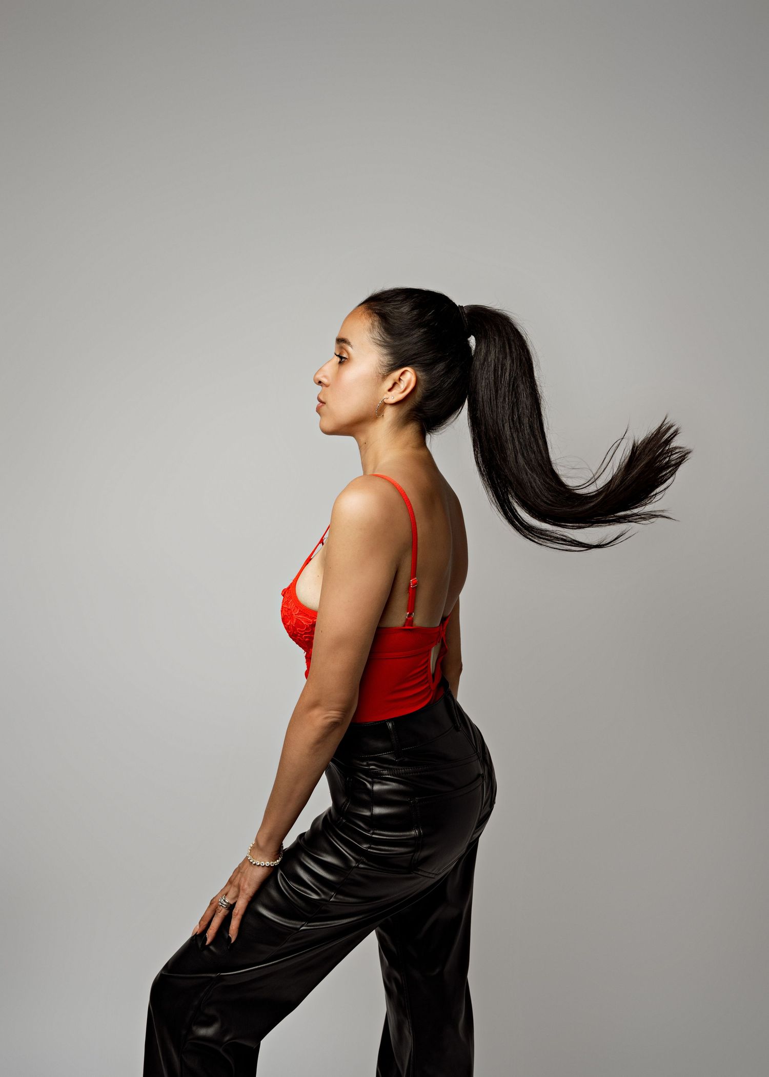 A dancer in a red top and black leather pants poses expressively against a neutral gray background.
