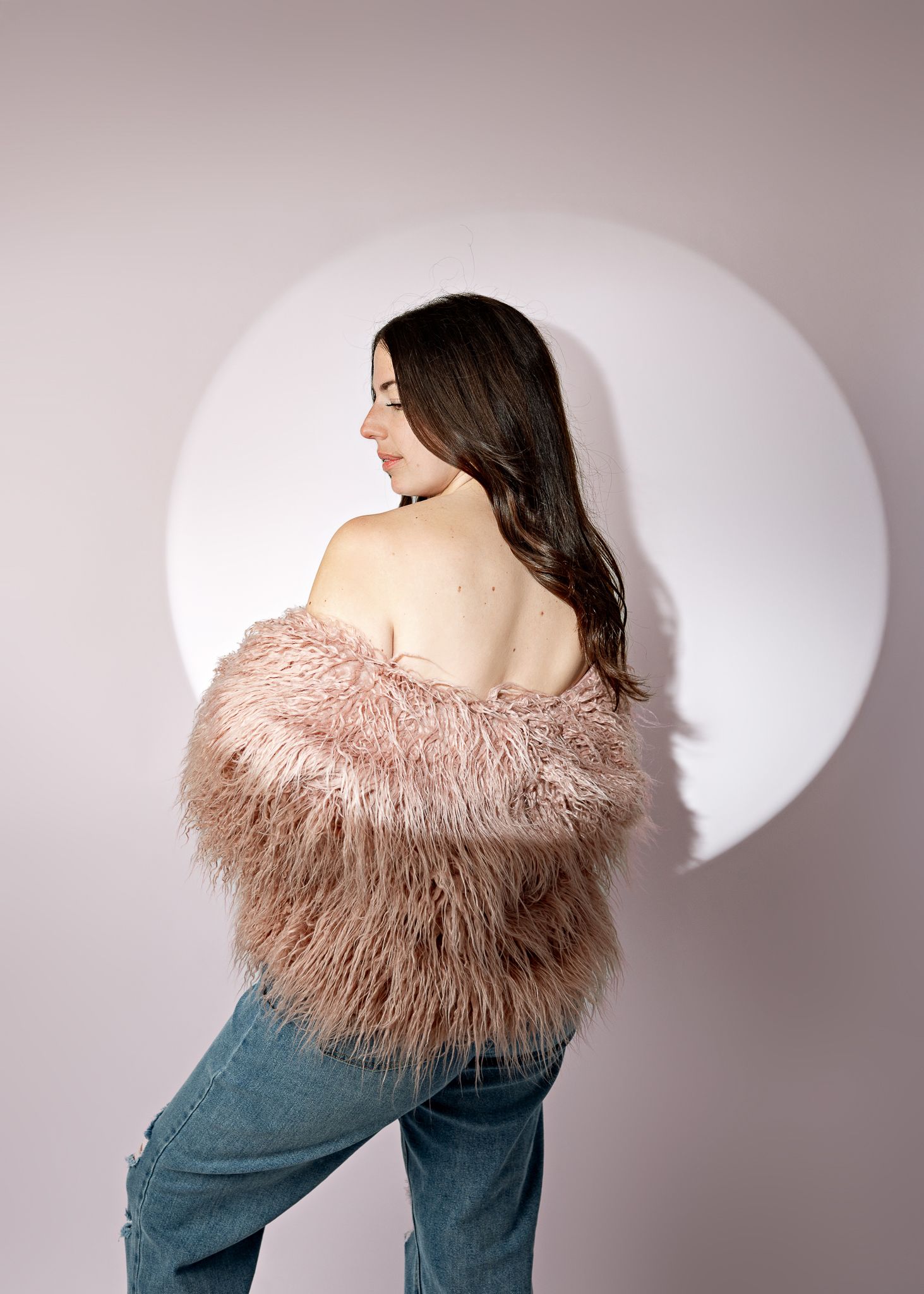 Model wearing pink fluffy faux fur coat and blue jeans poses against white wall with circular backdrop.