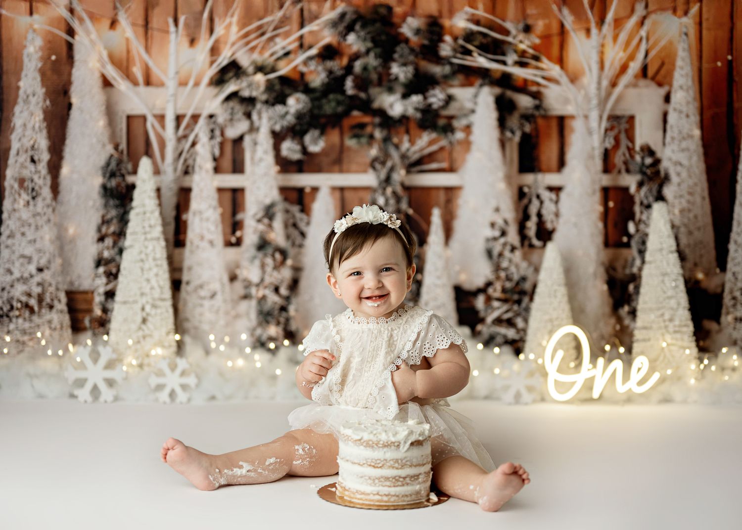 Festive winter wonderland cake smash photo setup with illuminated decorative cone trees and neon ONE sign.