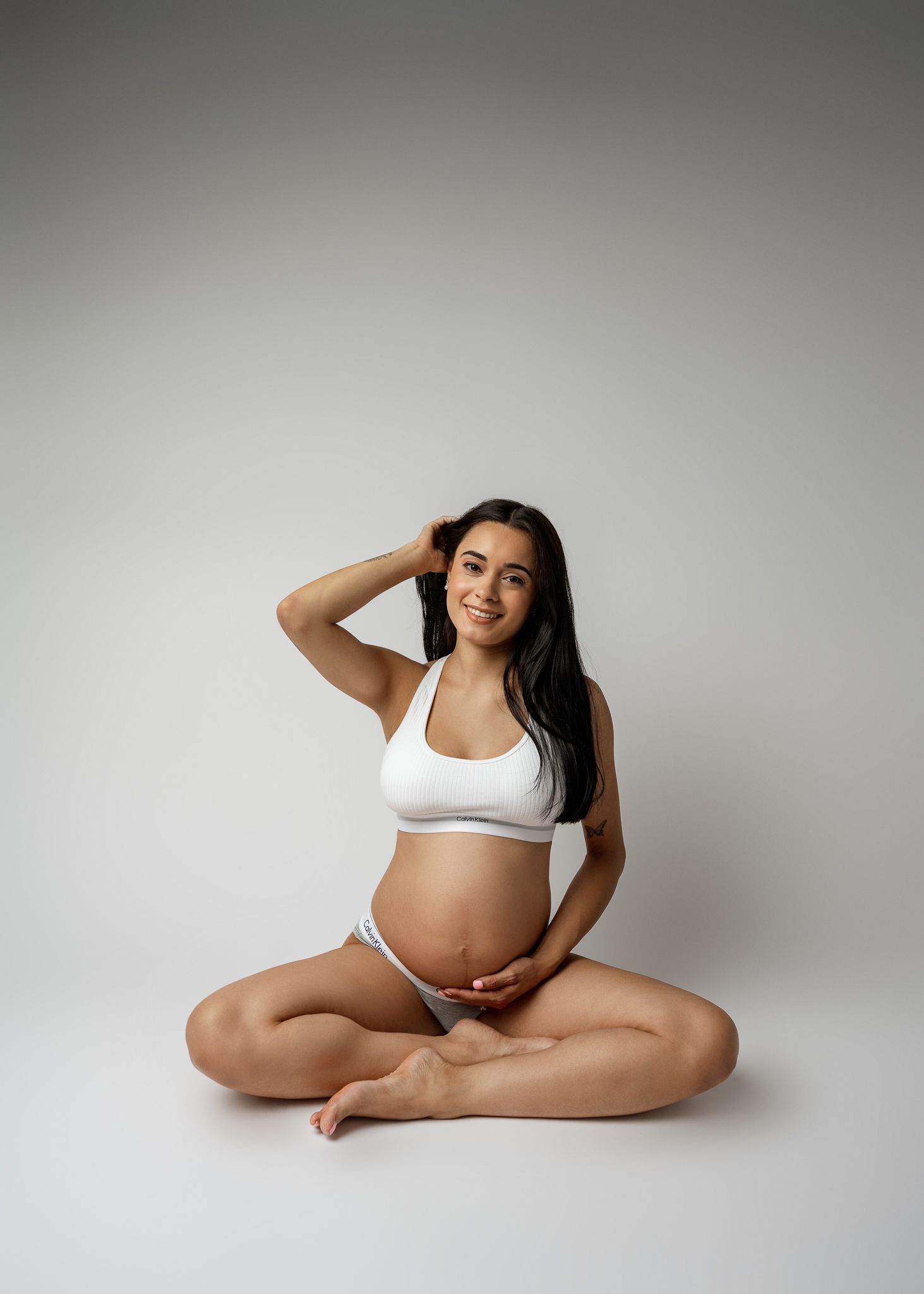 Maternity photo series showing a pregnant woman in white underwear posing gracefully against a light gray backdrop.