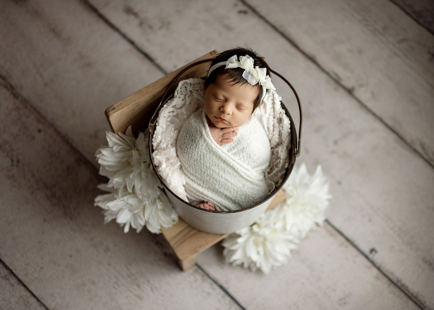 A series of newborn photography shots showing a sleeping baby in white against a rustic wooden floor with white flowers.