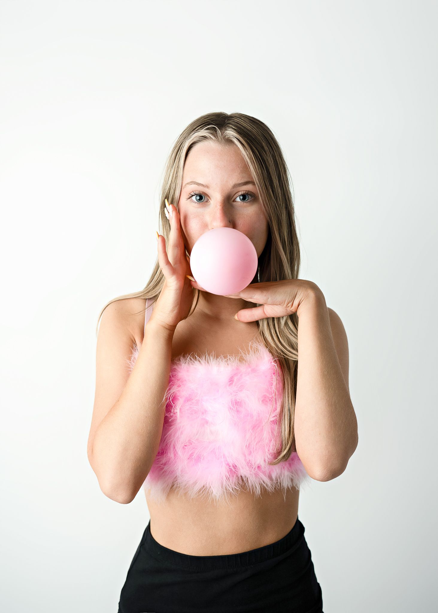 Person wearing pink fuzzy crop top blowing pink bubble gum against white background.