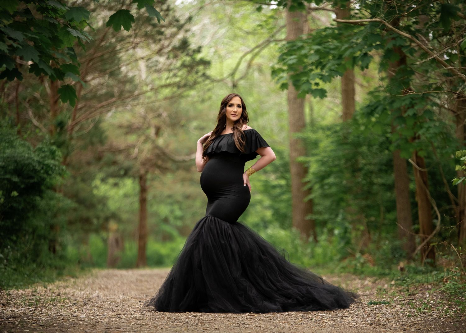 Stylish maternity photo featuring an elegant black tulle gown on a serene forest path surrounded by green trees.