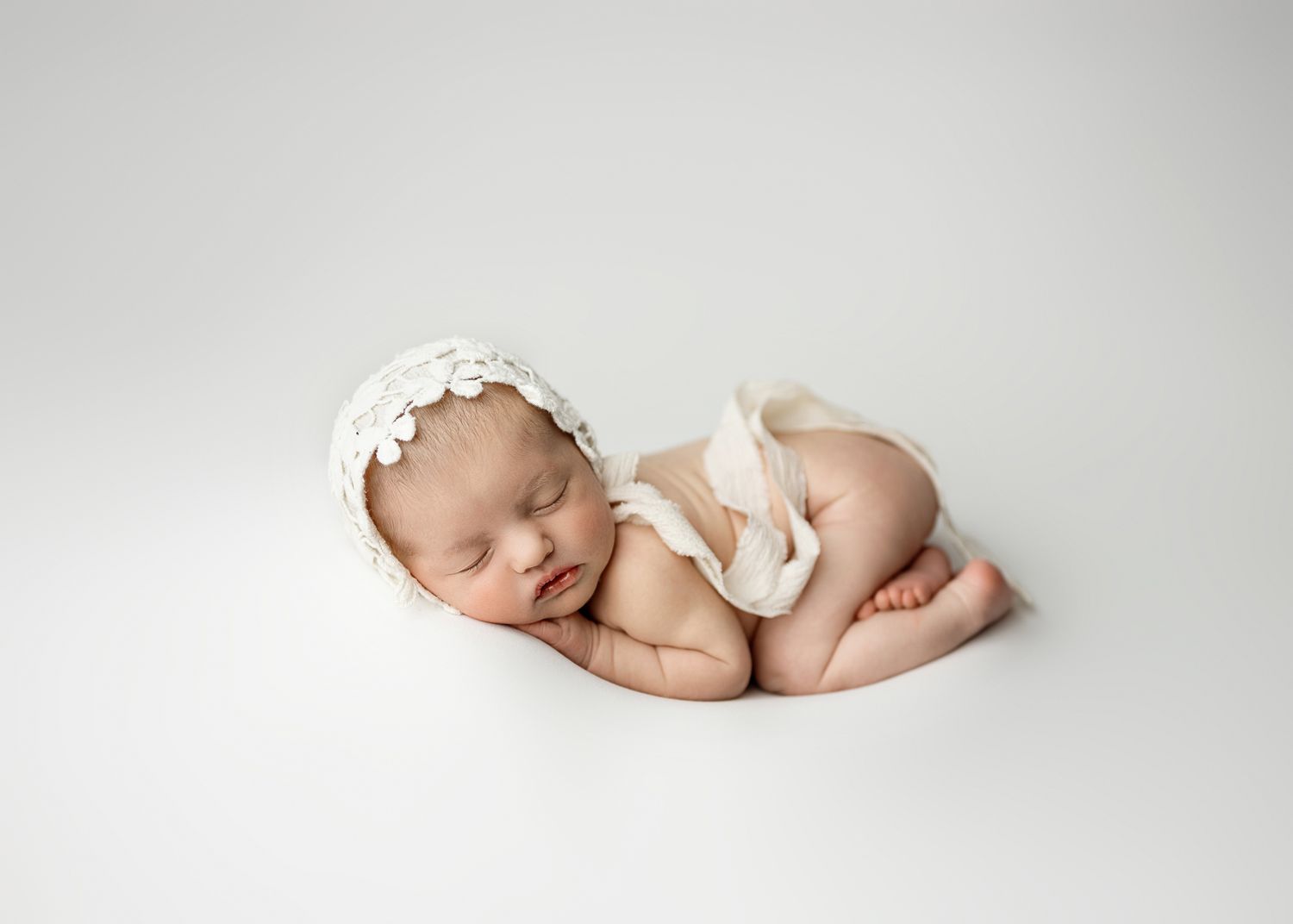 A sleeping newborn baby lies peacefully in white swaddling cloth against a soft white background.