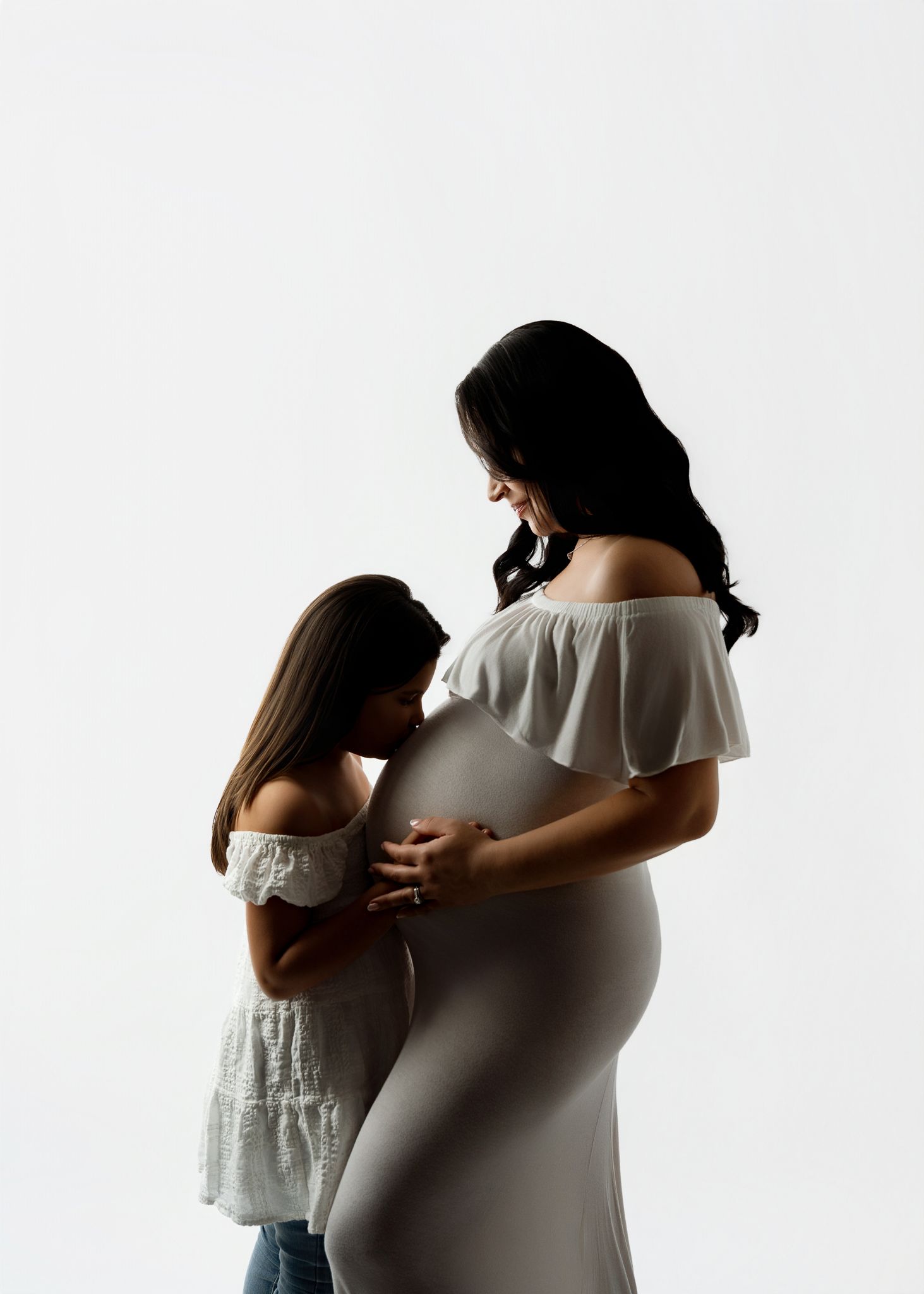 Artistic silhouette of pregnant woman in white dress with young child touching her belly against light background.