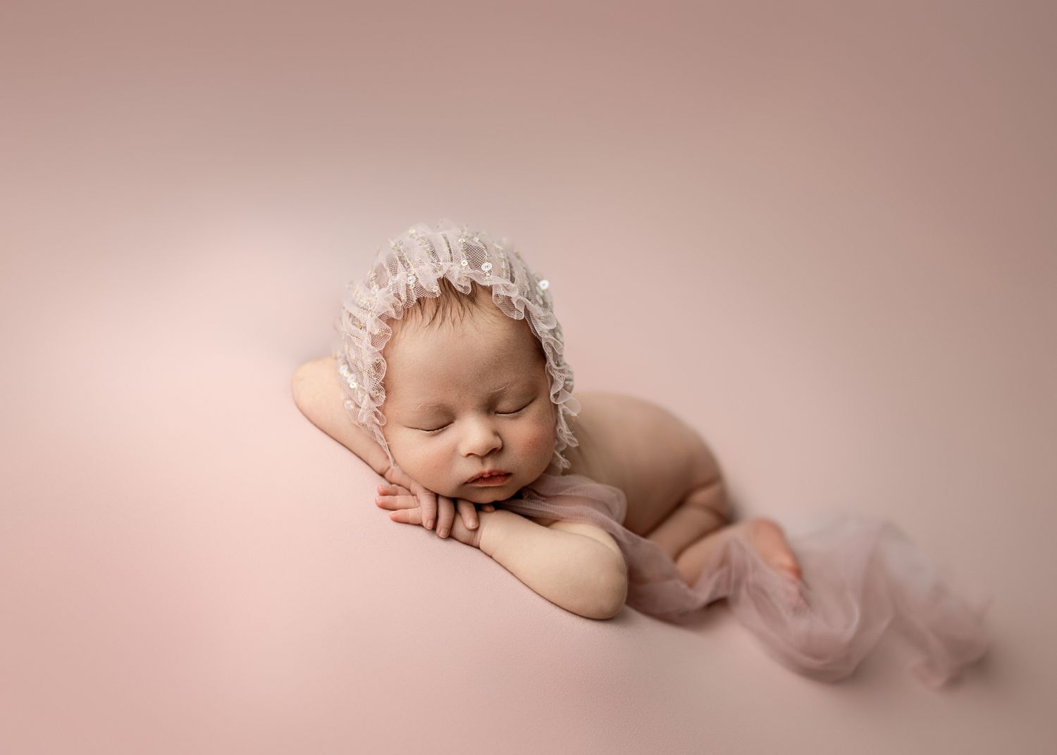 Sleeping newborn wearing lace bonnet resting on pink backdrop in artistic pose.