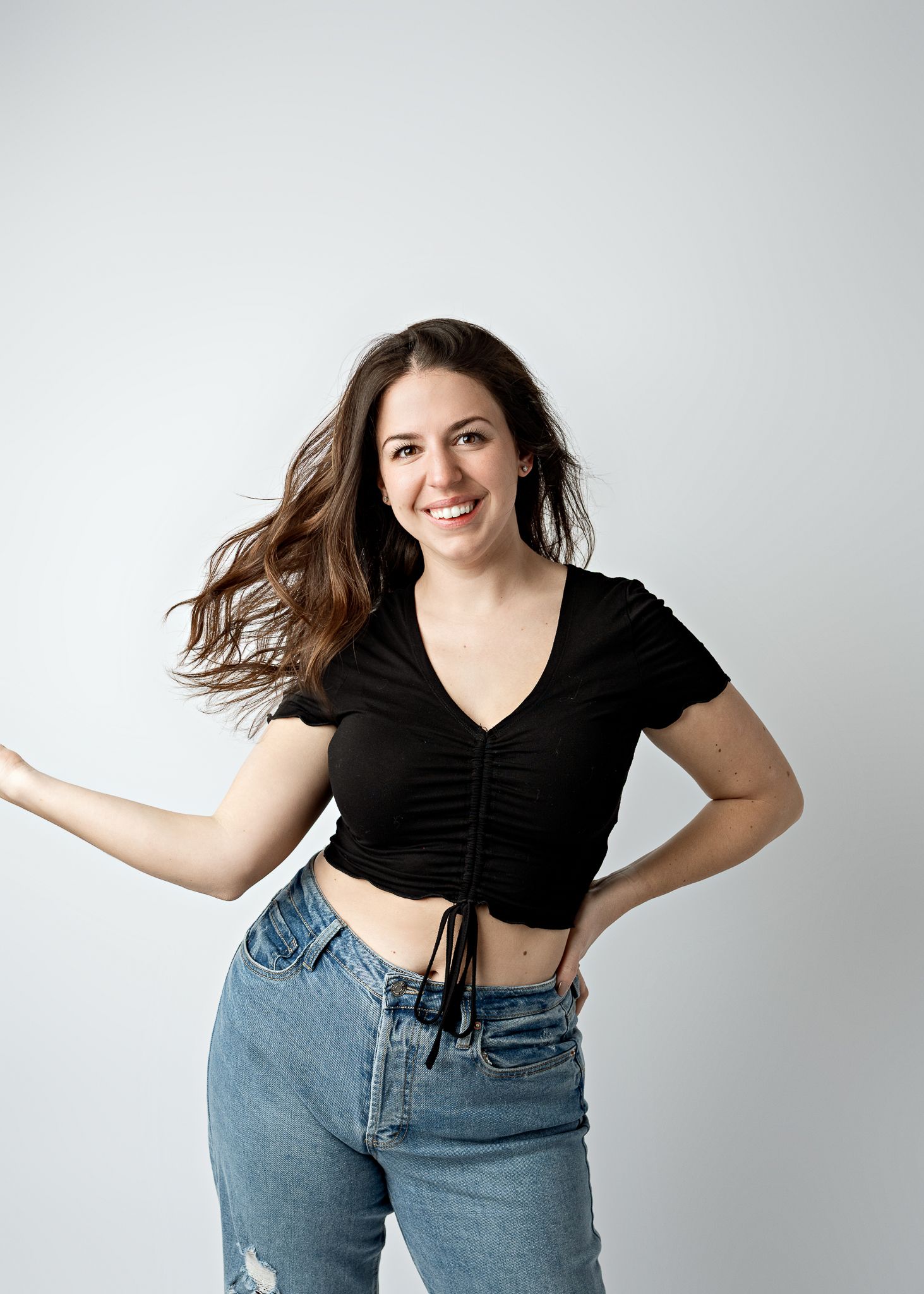 Person in black crop top and light blue jeans poses playfully against white background with windswept hair.