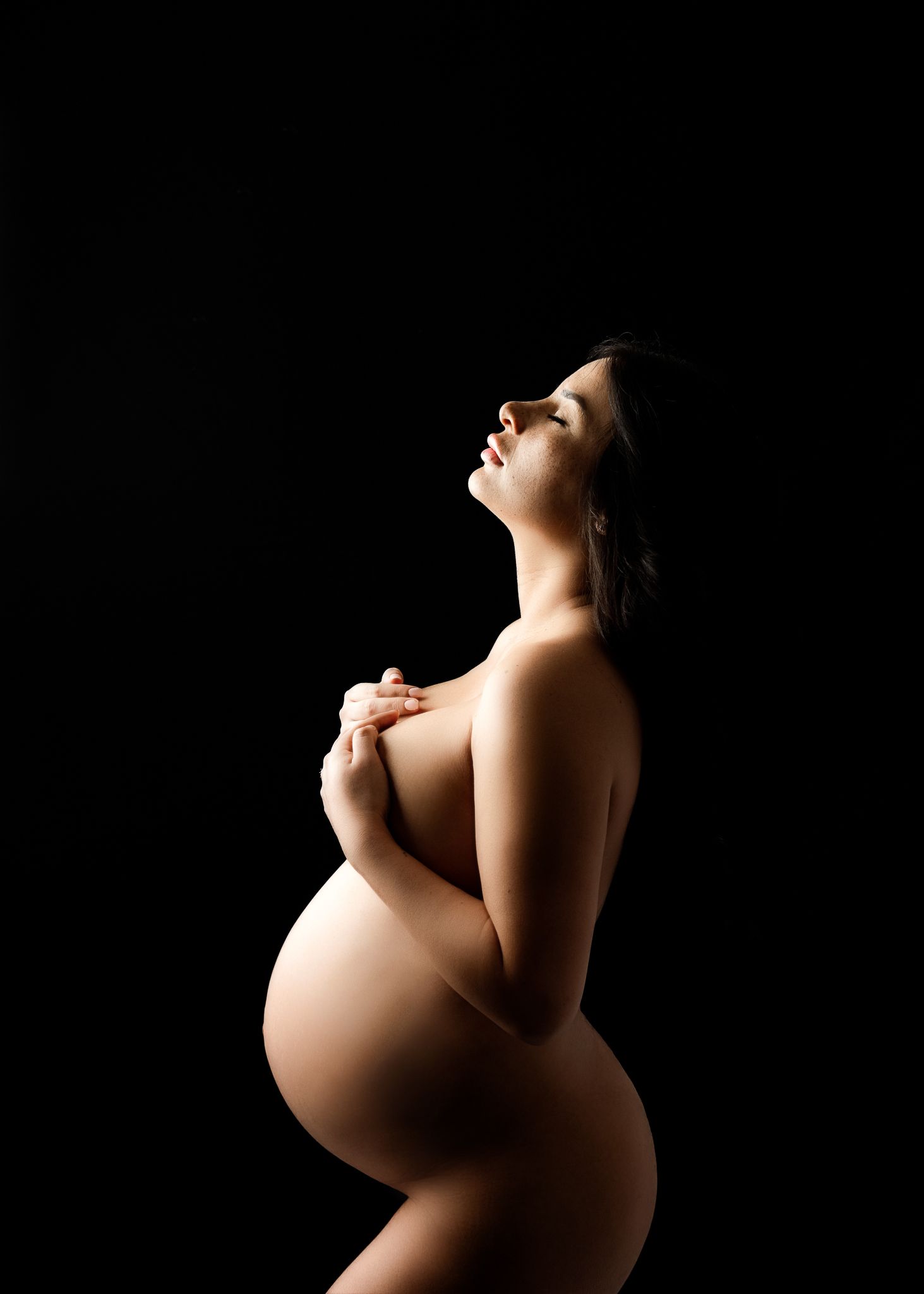 Artistic silhouette of an expectant mother in dramatic lighting against dark background, captured in profile view.