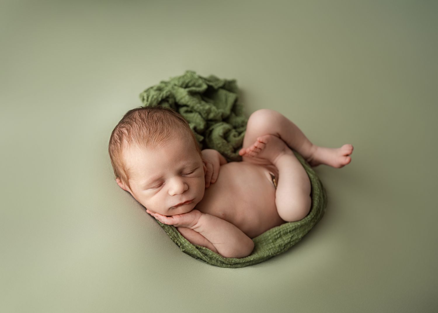 Peaceful sleeping newborn nestled on soft sage green fabric in minimalist portrait setting.