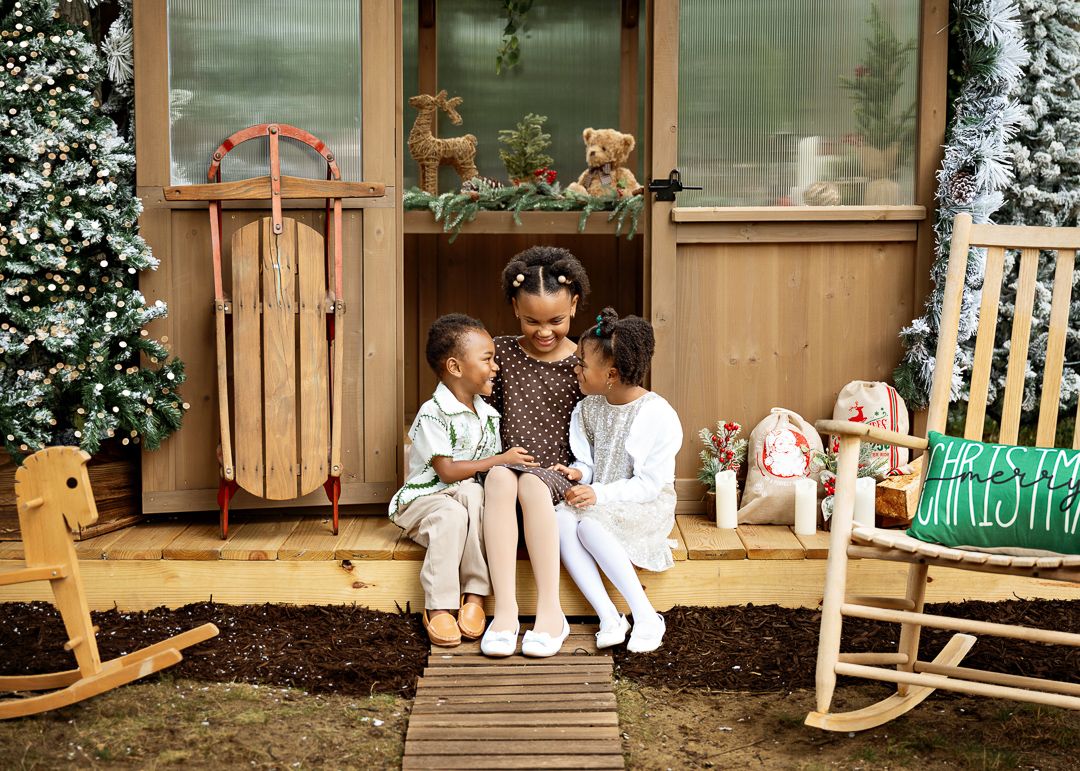 Three Black children sit on a wooden porch surrounded by Christmas decorations, sleds, and rocking chairs.
