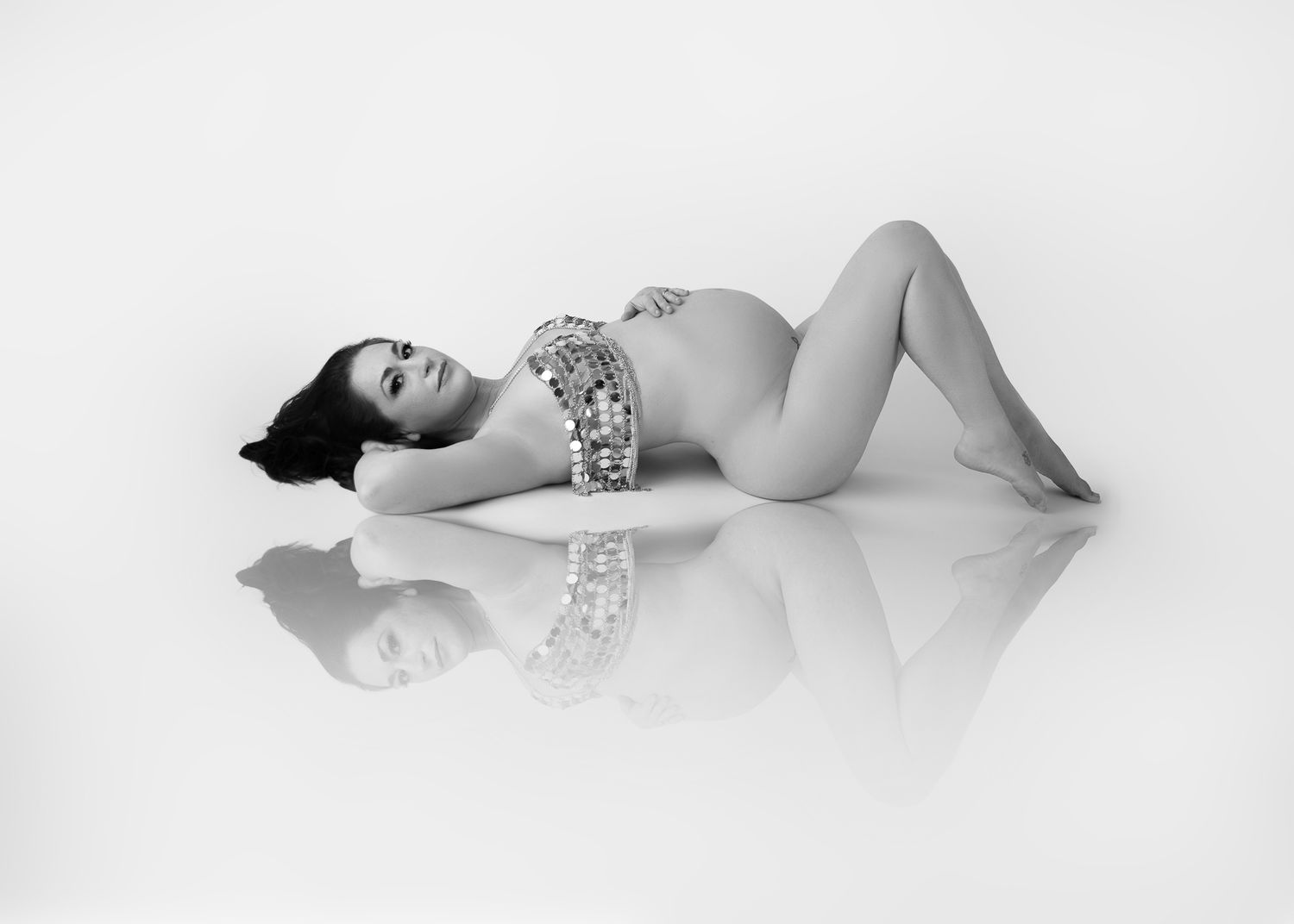 Black and white artistic maternity portrait showing a figure in a floral bodysuit reclining gracefully on a reflective surface.