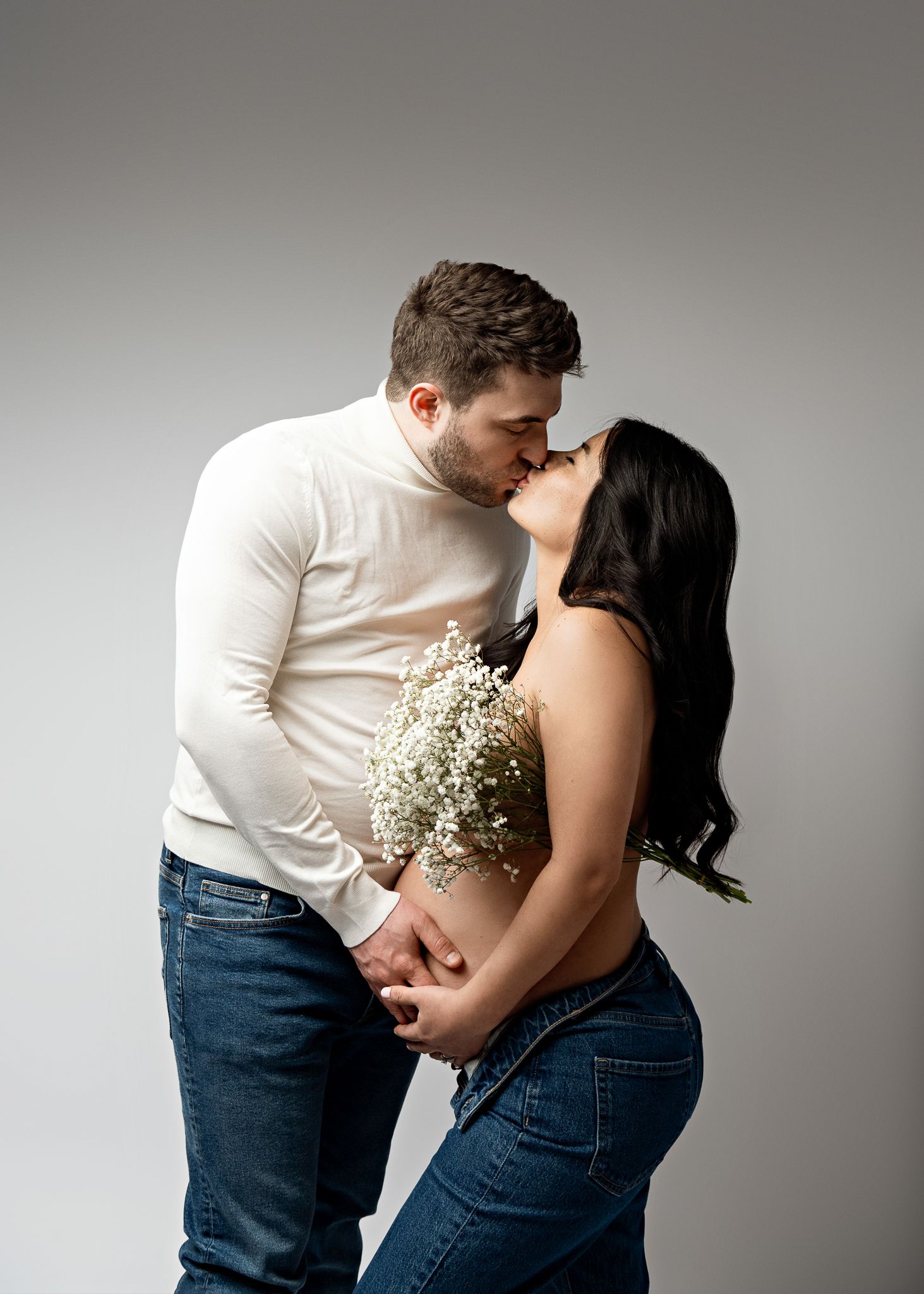A romantic couple share an intimate moment while holding delicate white flowers in a minimalist studio setting.