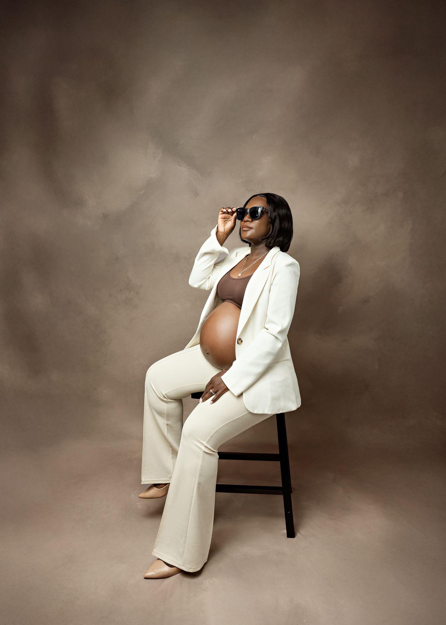Stylish pregnant person in all-white business suit and sunglasses poses confidently on wooden chair against brown backdrop.