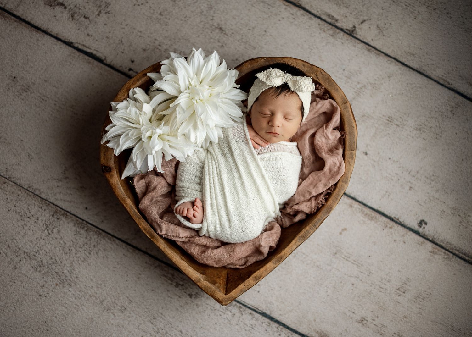 A sleeping newborn baby wrapped in white fabric lies peacefully in a rustic wooden bowl on weathered wooden floorboards.