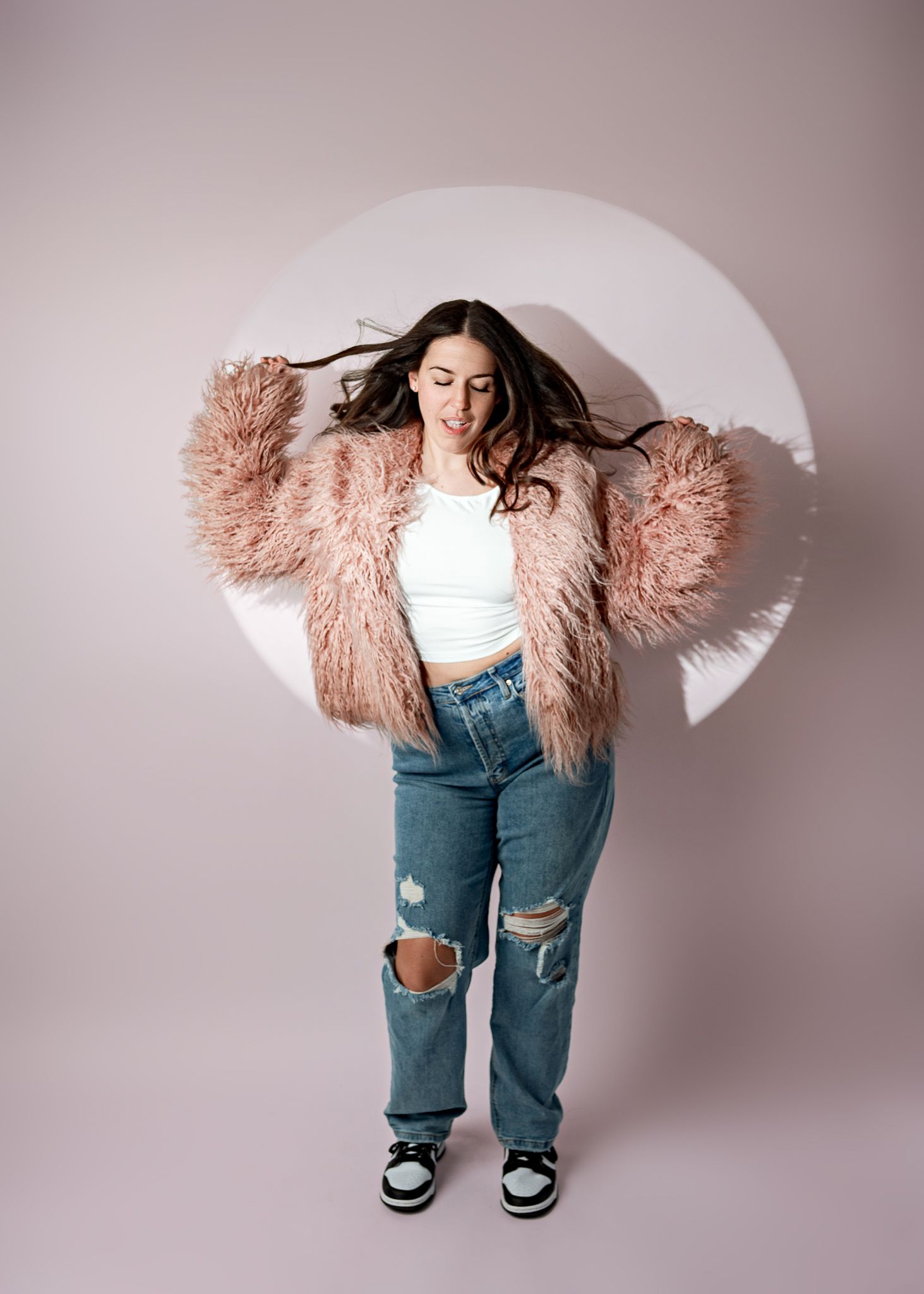 Trendy outfit featuring a pink fluffy faux fur coat, white crop top, distressed jeans and sneakers against a white backdrop.