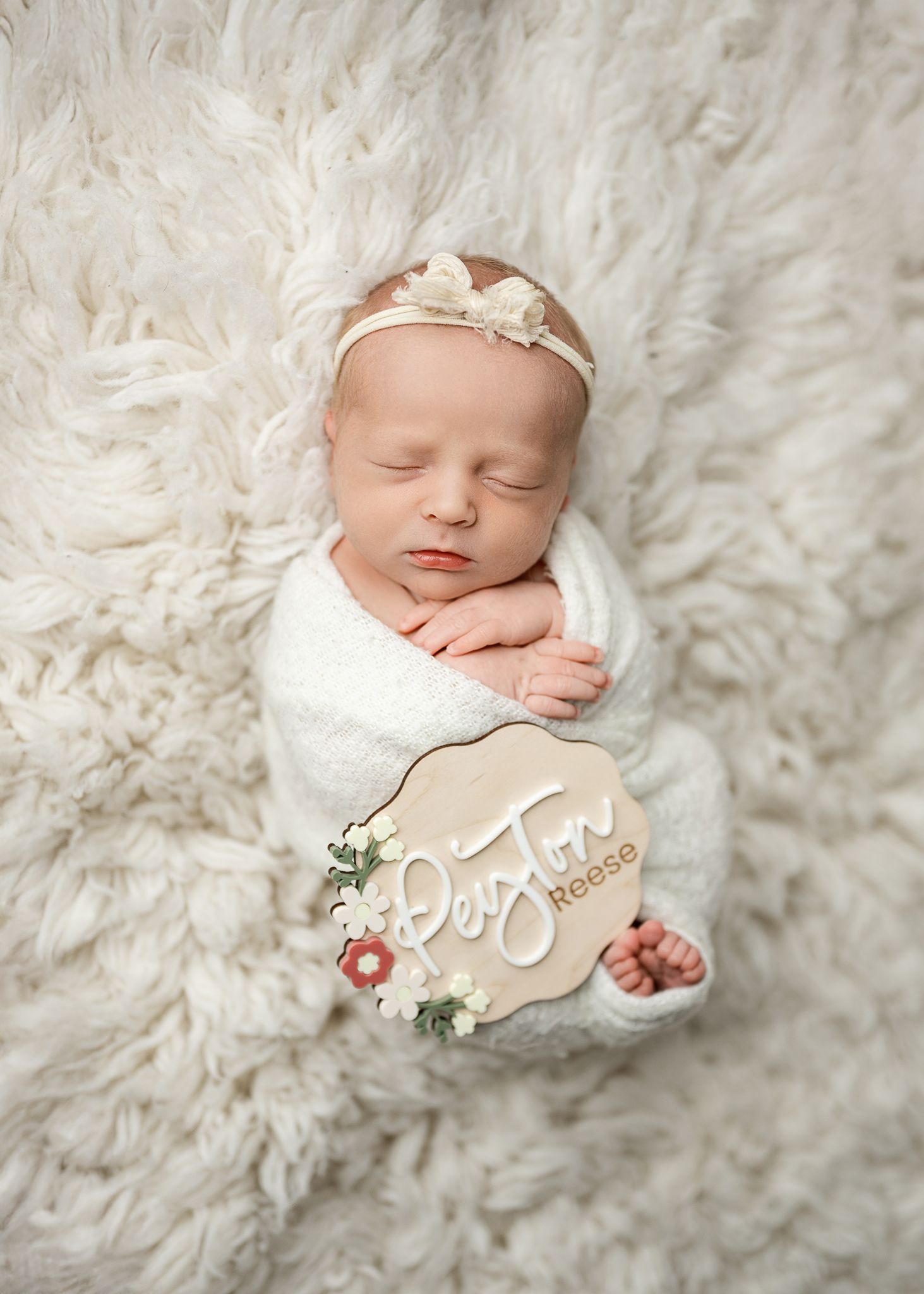 Newborn baby sleeping peacefully on white fluffy fur blanket wearing delicate headband and floral swaddle wrap.