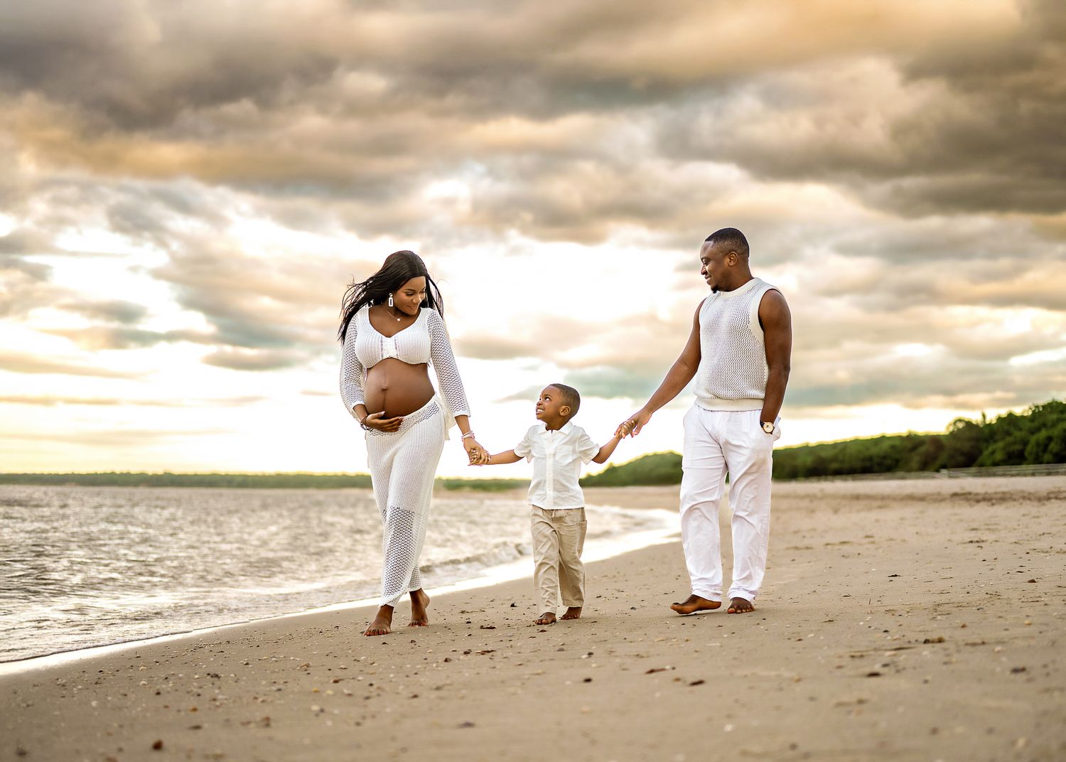 Family in white clothing holds hands while walking on beach at sunset with dramatic cloudy sky over ocean.