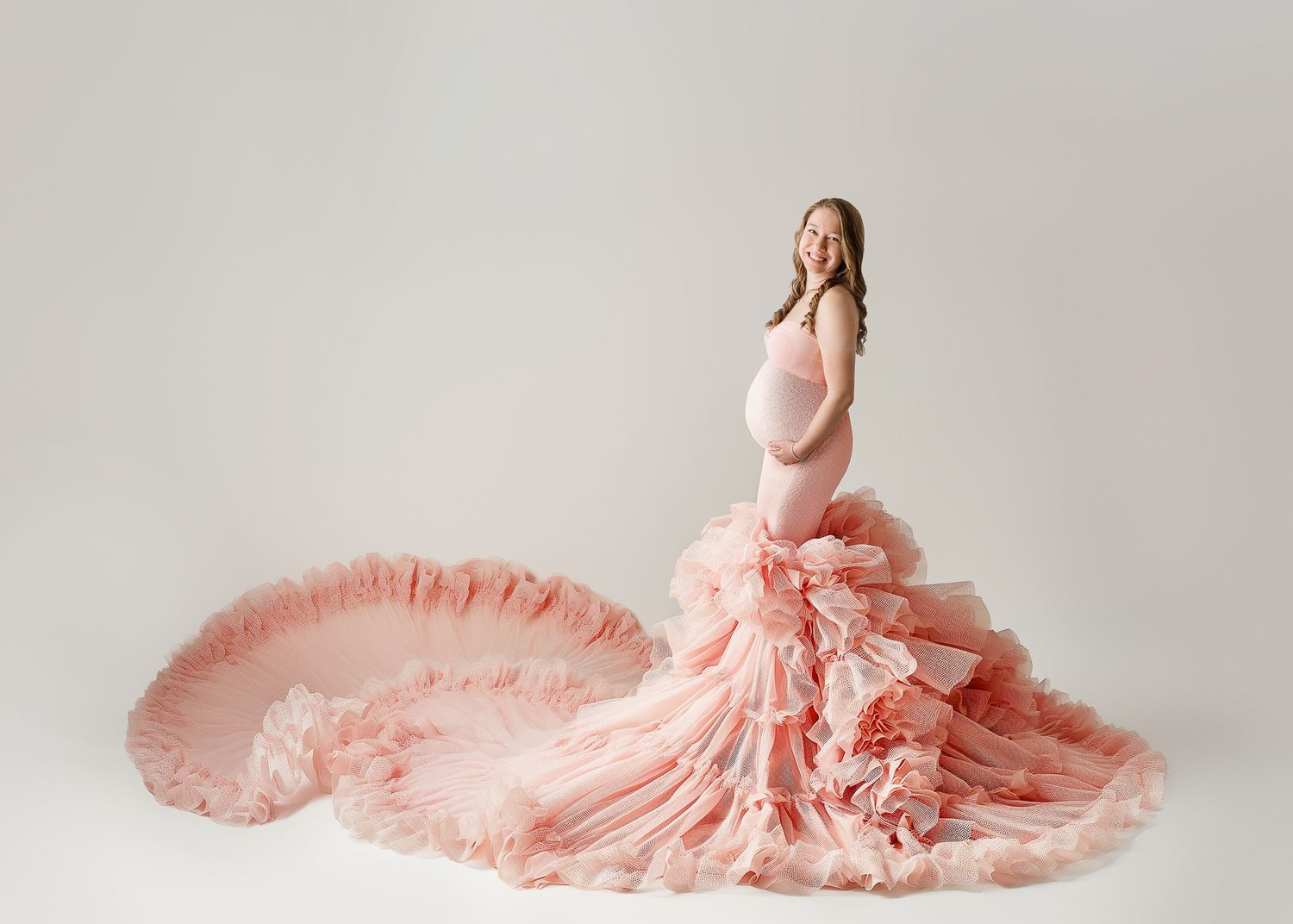 A pregnant figure poses in an ethereal peach-colored gown with a dramatic ruffled train against a light background.