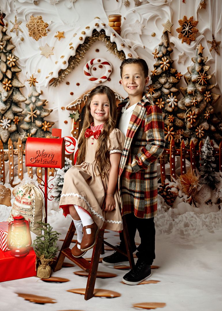 Two children pose in festive holiday outfits before a Christmas backdrop with trees, candy canes, and a red mailbox.