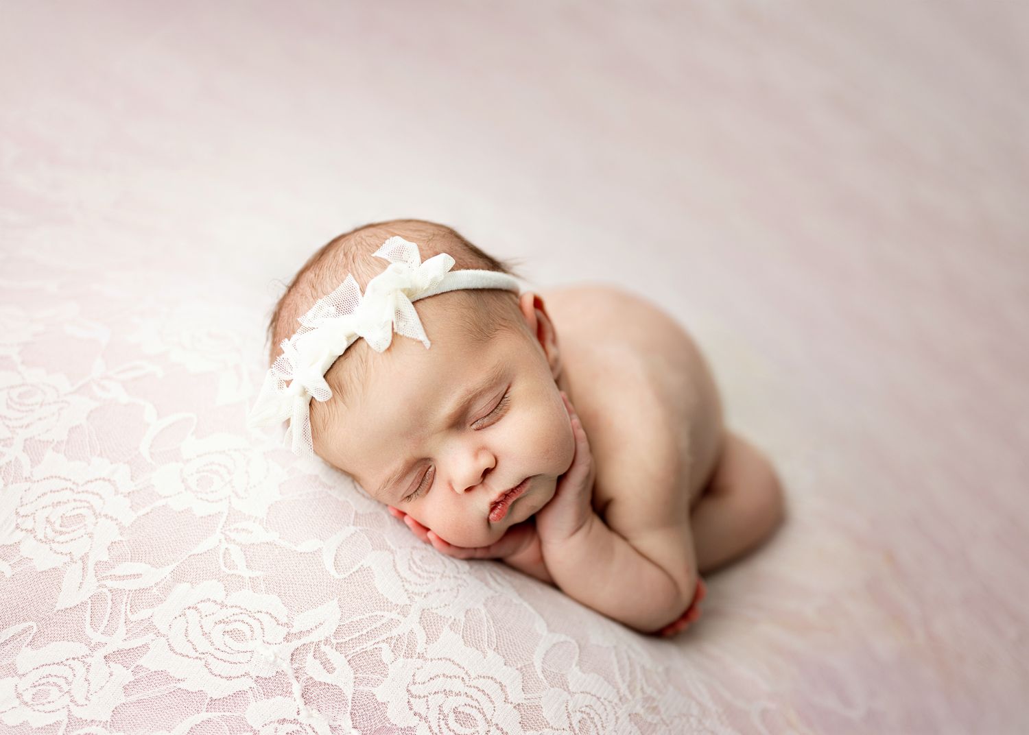 Sleeping newborn baby wearing white headband rests peacefully on soft pink lace blanket.