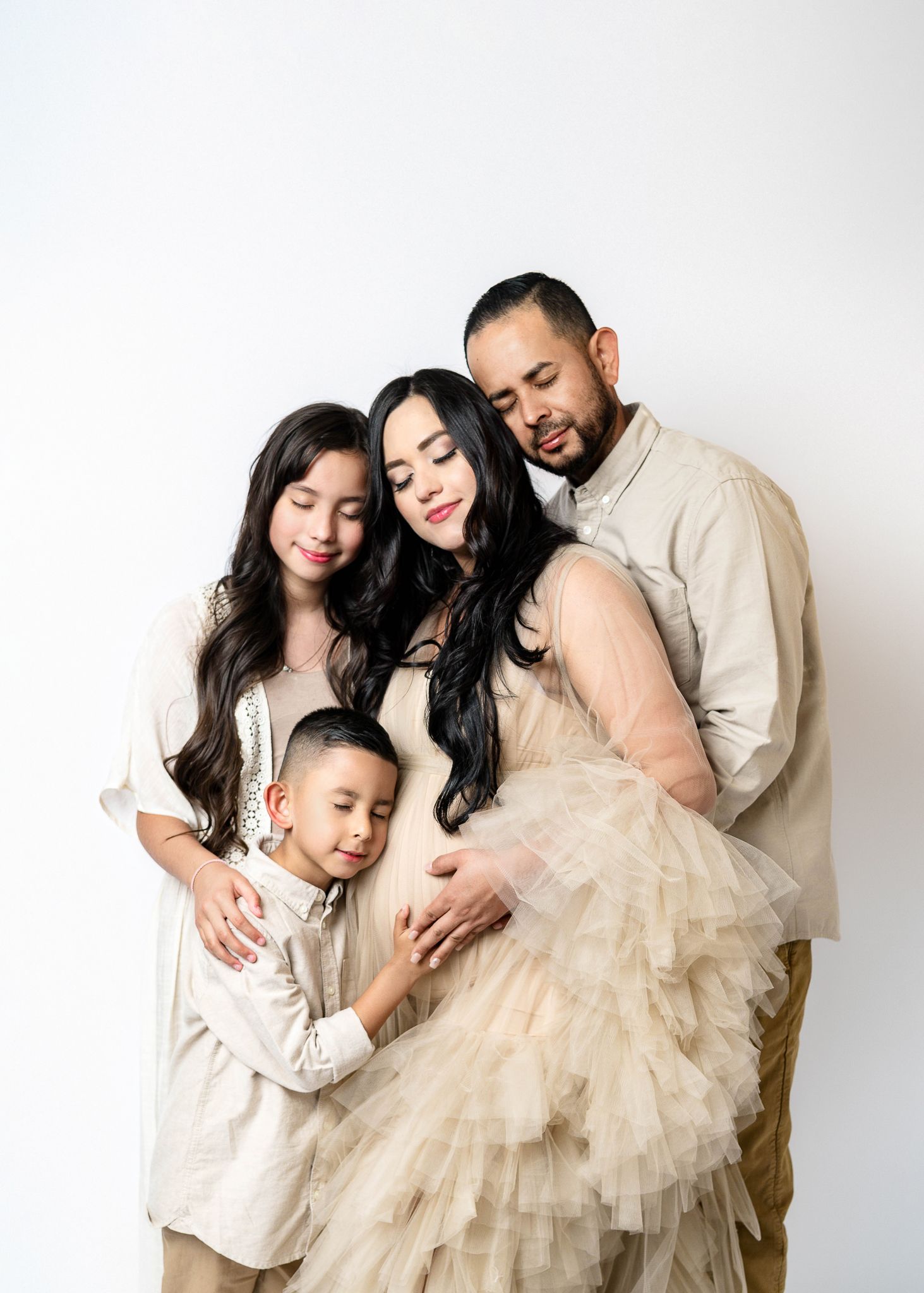Family gathered in white studio wearing neutral colors for maternity photoshoot.